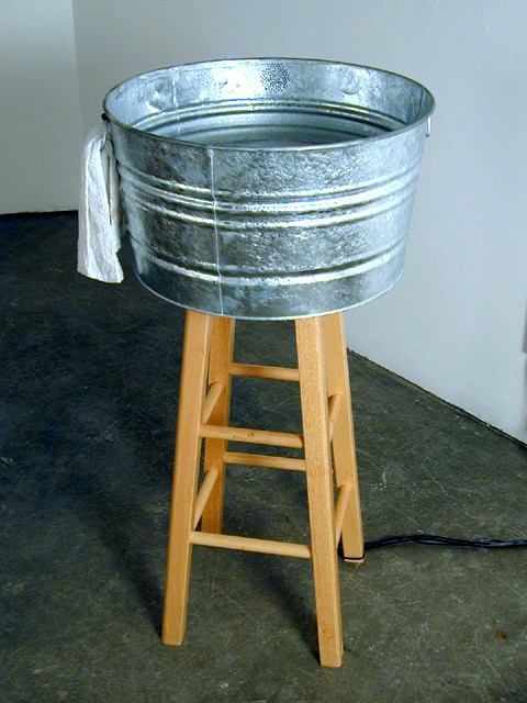  Ablution 20” x 20” x 40”, Galvanized washing tub, water heater, soap suds, towel, photo sensor, video, sound :: 2002.  Visitors drawn to the water by its warmth and fragrant suds, trigger a video loop that is projected onto its surface. This only ha