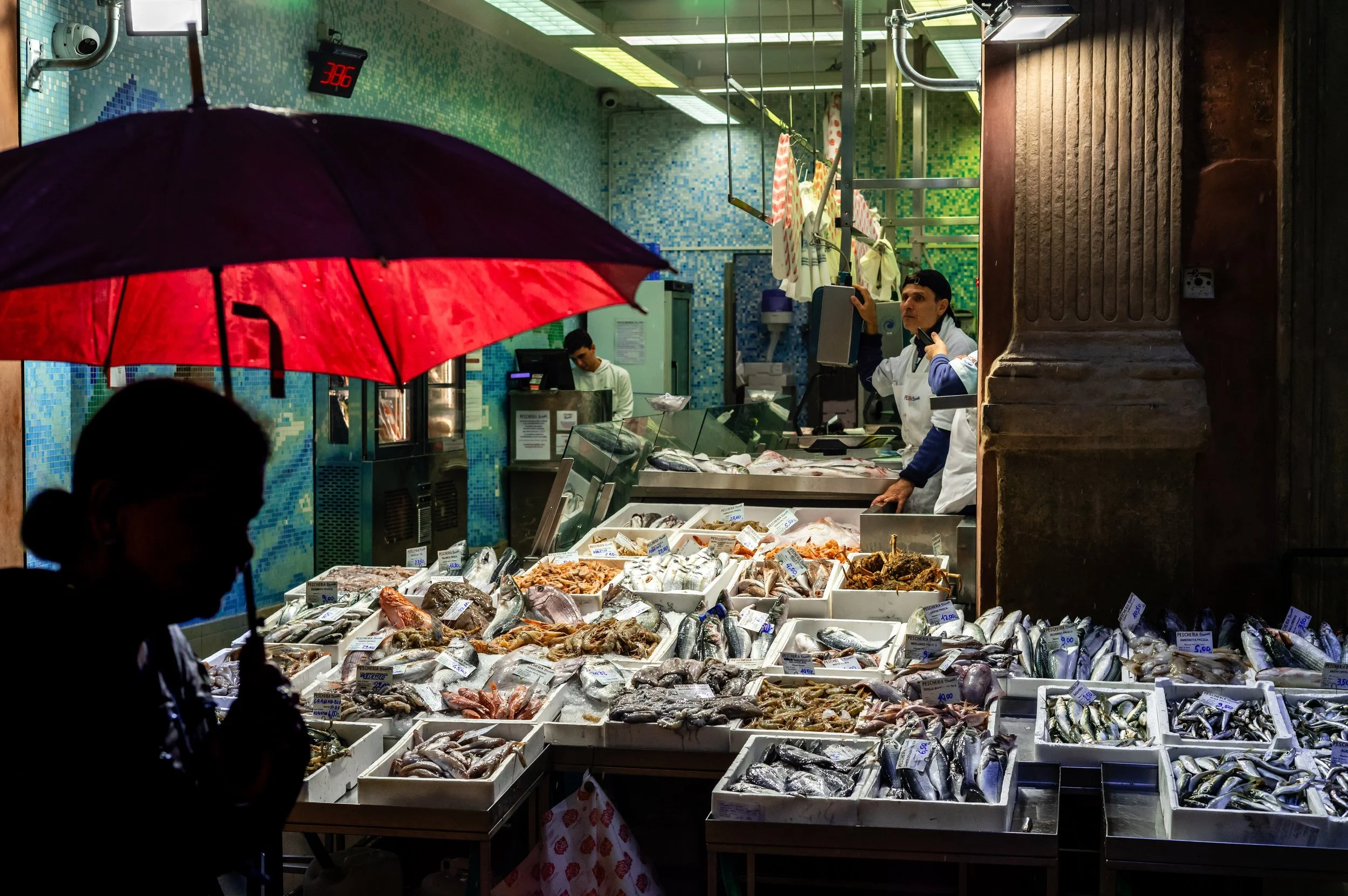 A fish market stall displaying various fresh fish and seafood for sale, with vendors behind the counter and a person holding a red and black umbrella in the foreground. Bologna, Italy.