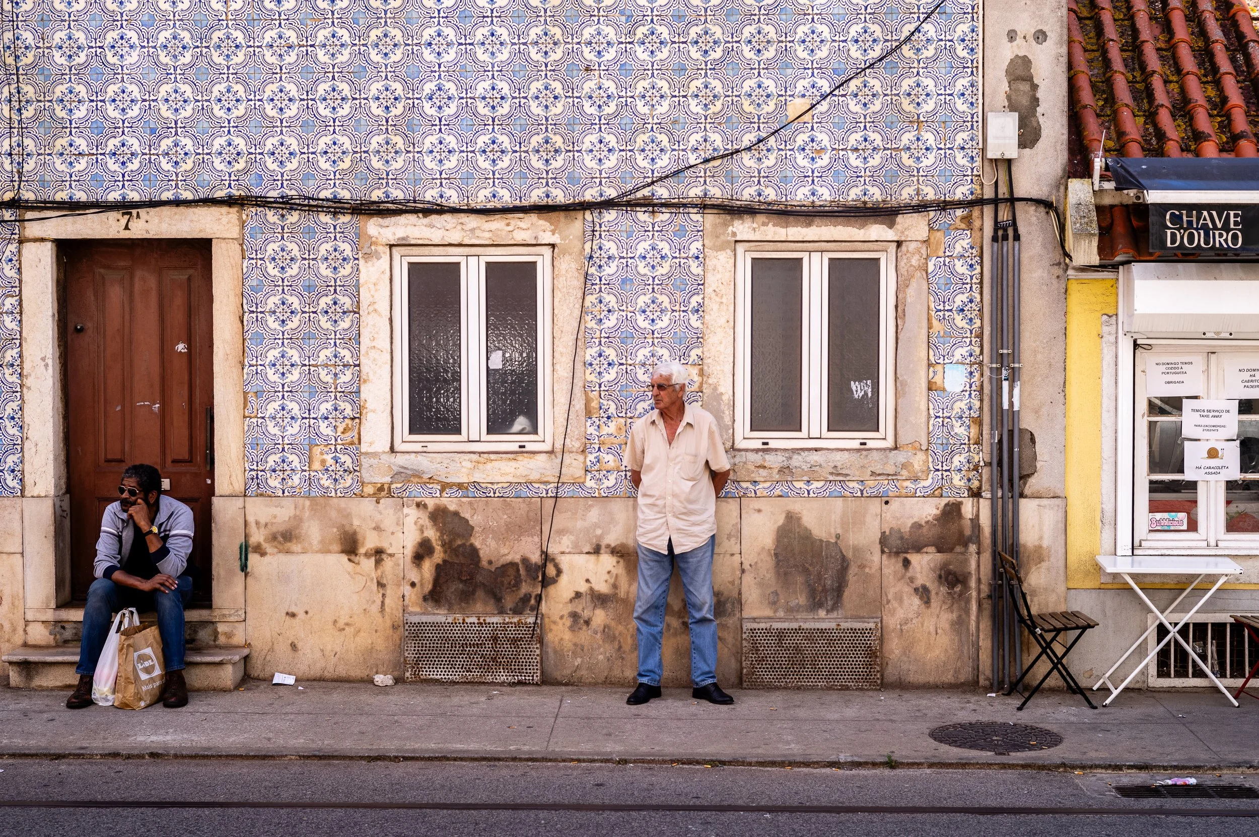 Street scene with two men, one sitting on steps near a door and the other standing in front of a wall with decorative tiles, windows, and electrical wiring. Lisbon, Portugal.
