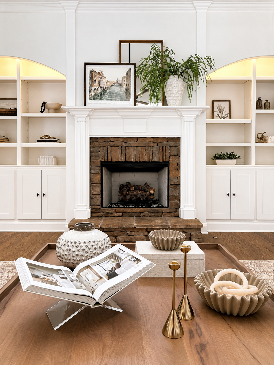 Living room with white built-in shelves, a brick fireplace with a white mantel, and decorative items on the table and shelves, including a large white vase with green foliage, framed pictures, and gold candle holders.