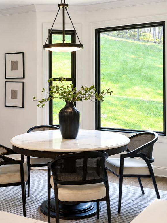 A round white marble dining table with a black vase holding green foliage, surrounded by black and white chairs, light fixture hanging above, and windows showing a grassy outdoor scene.