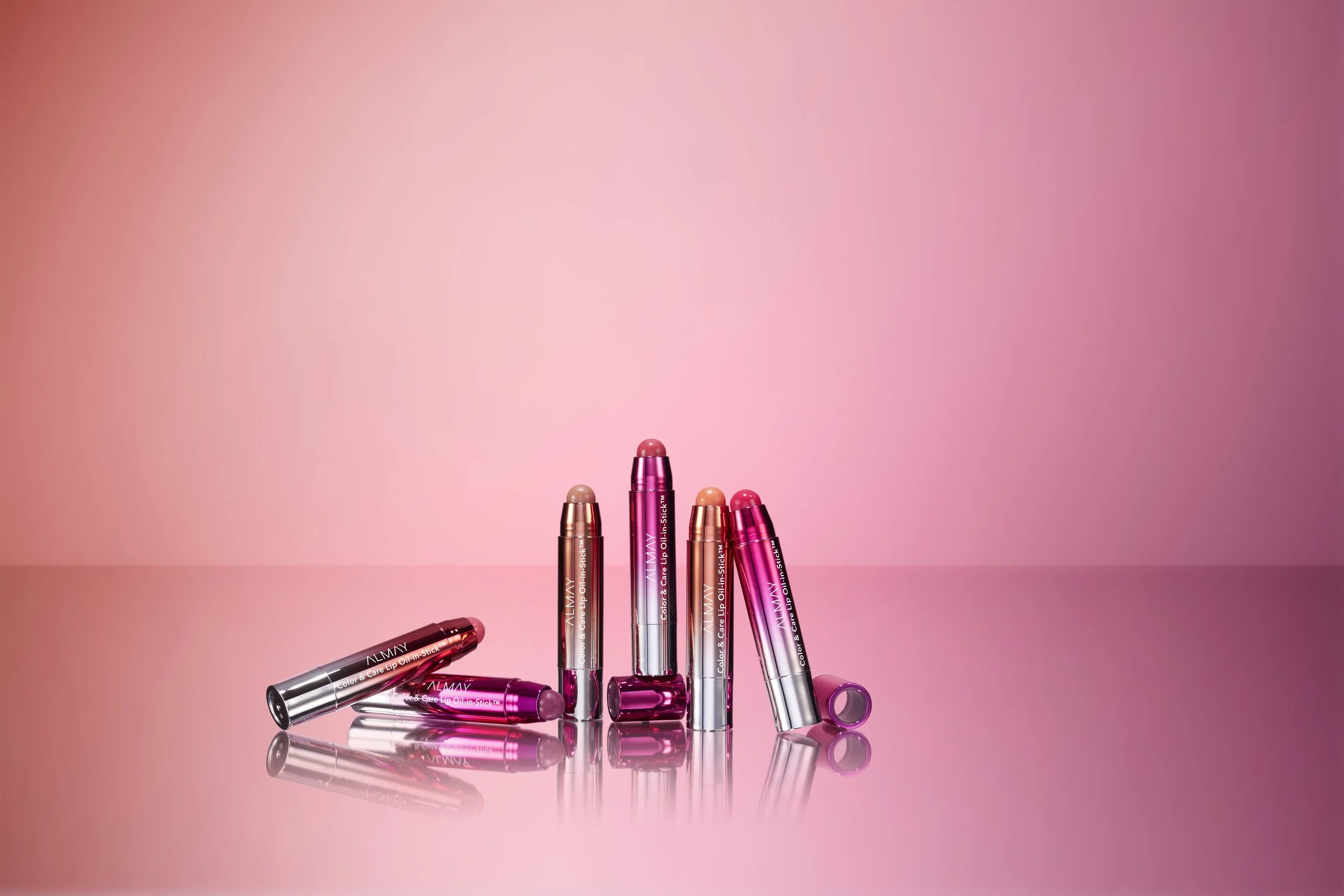 Several lip balm sticks in shades of pink, coral, and nude arranged on a pink background, with some lying down and one standing upright.