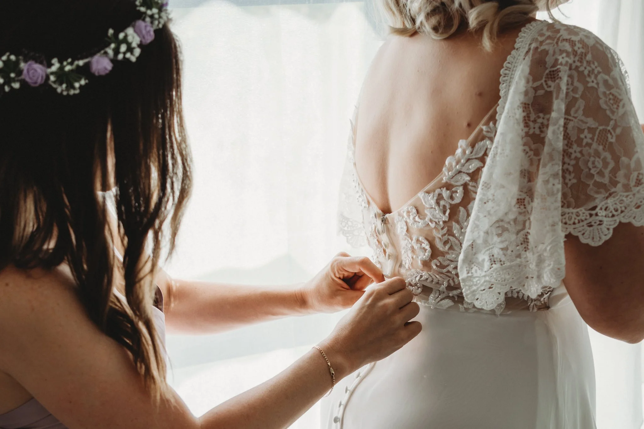 A woman helping a bride button her wedding dress. The bride's dress is lace and has intricate embroidery. The woman has long, wavy brown hair and is wearing a bracelet.