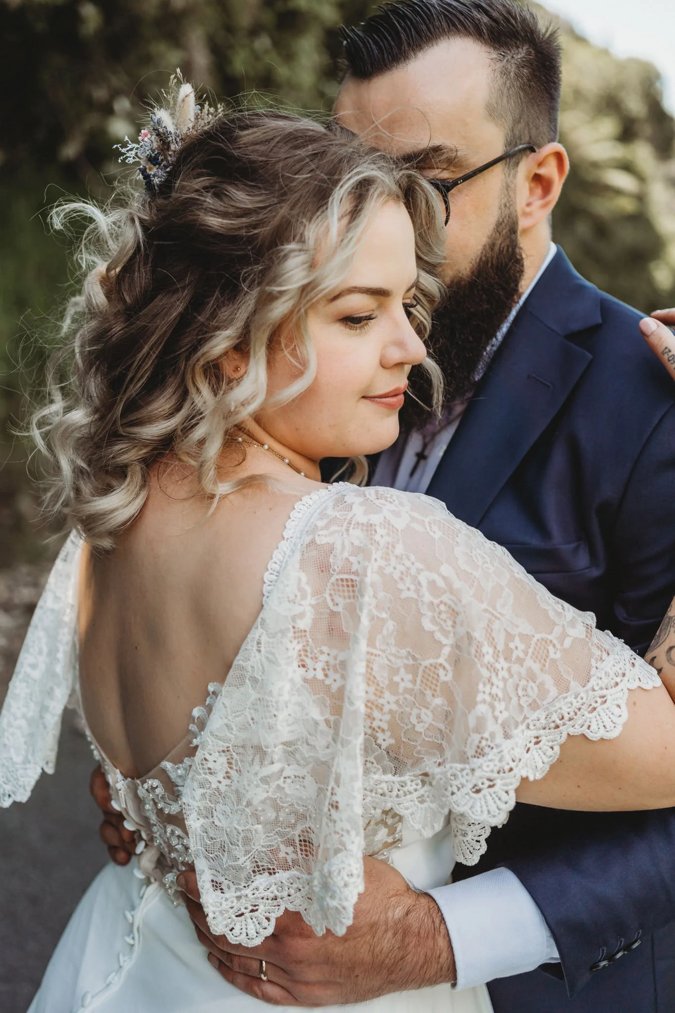 A bride in a wedding dress with lace detail and a groom in a navy suit sharing an intimate moment outdoors.