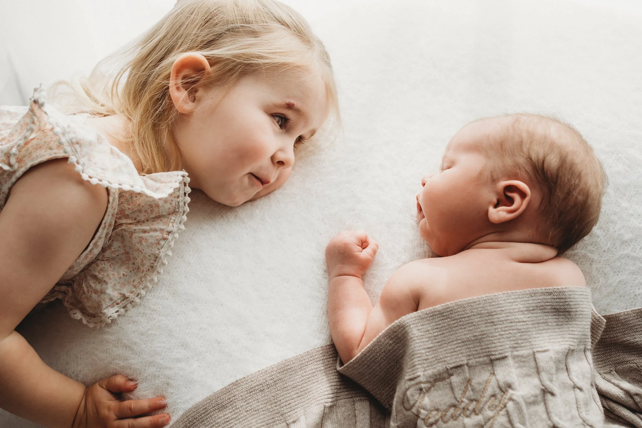 Big sister looks over at her newborn baby sister in this natural sibling moment photograph