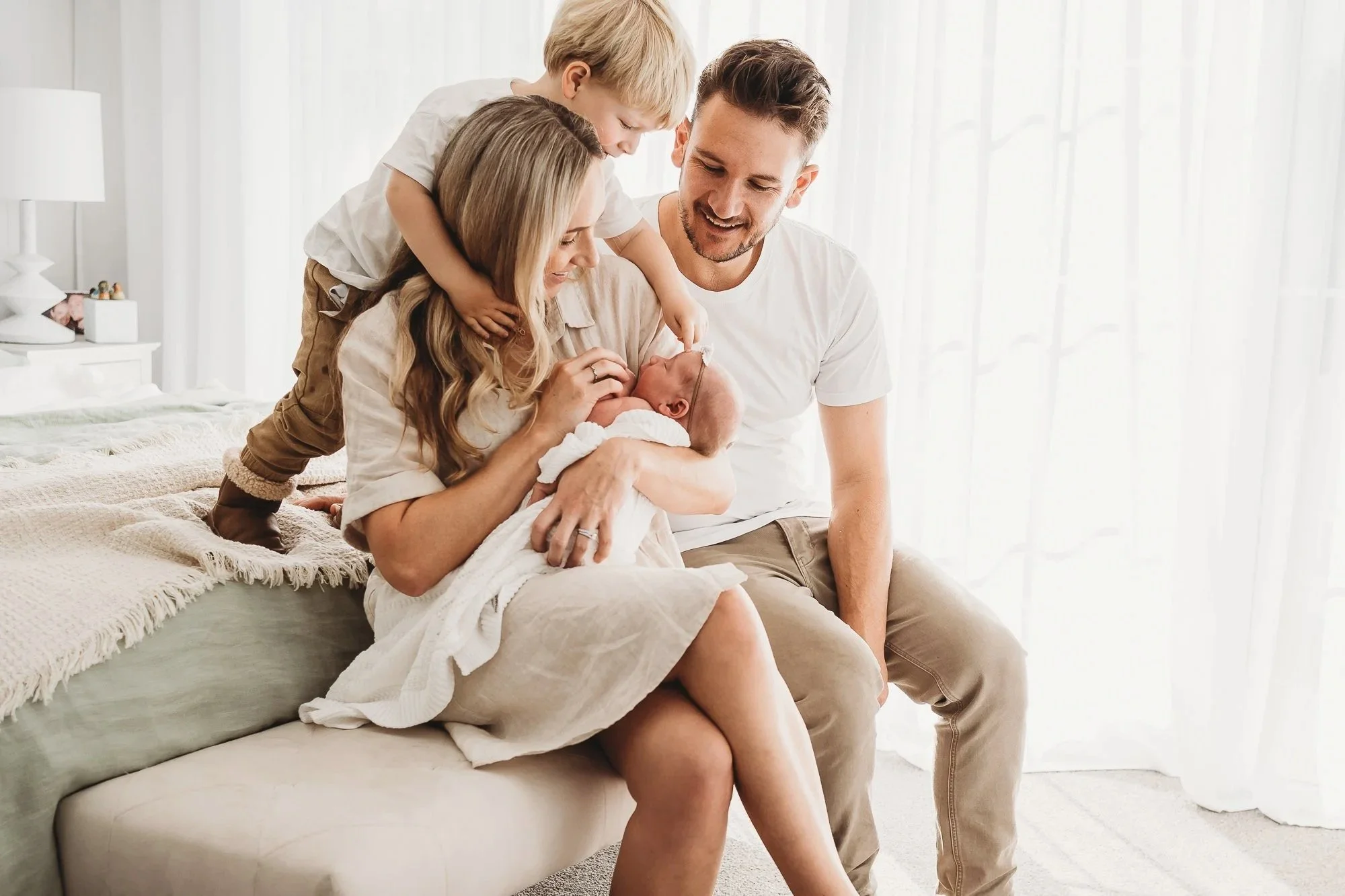 A family of four family sitting on a bed in a cozy, bright room. The mother is holding a newborn baby, while a young child leans over her shoulder. The father is sitting nearby, all sharing a joyful moment together.