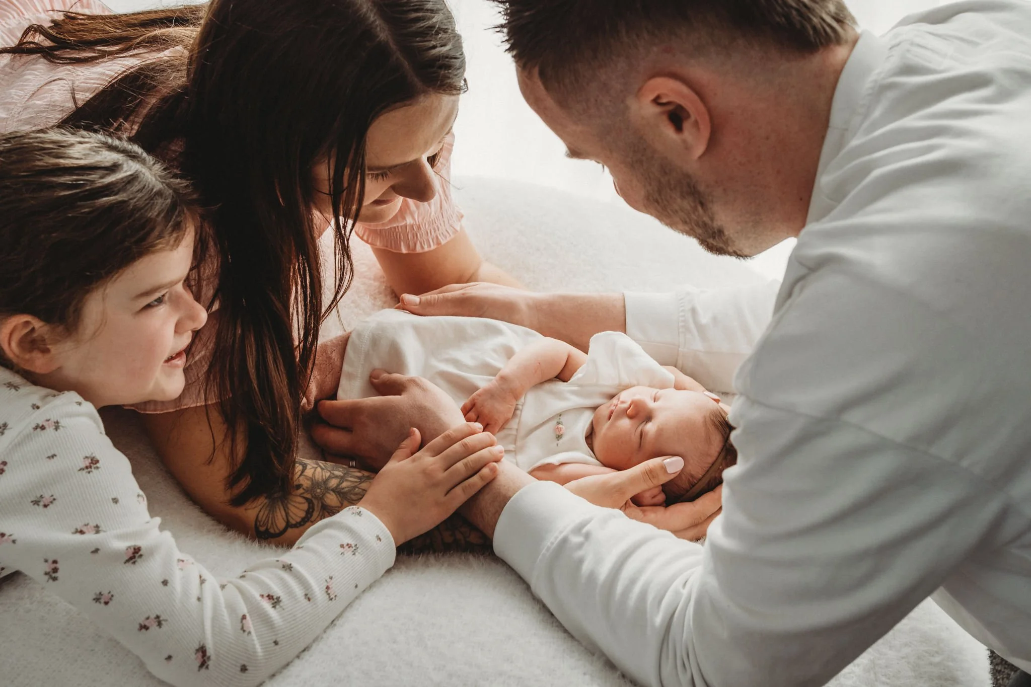 A family in a hospital room surrounding a newborn baby. The mother is lying on the bed with her head down, the father is leaning over the baby, and a young girl is holding and smiling at the baby. The scene is tender and joyful.