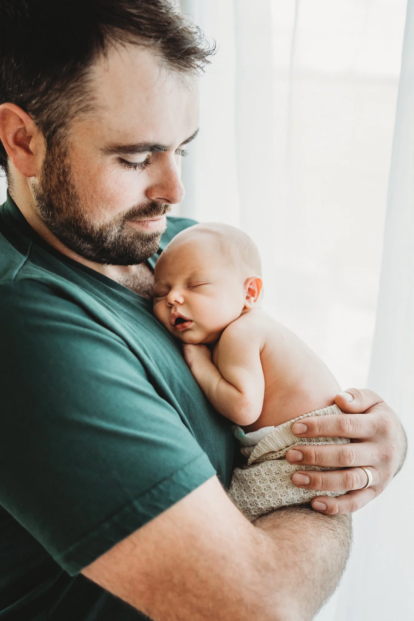 A man with a beard and brown hair holding a sleeping baby with light skin, light hair, and closed eyes, against a backdrop of white curtains.