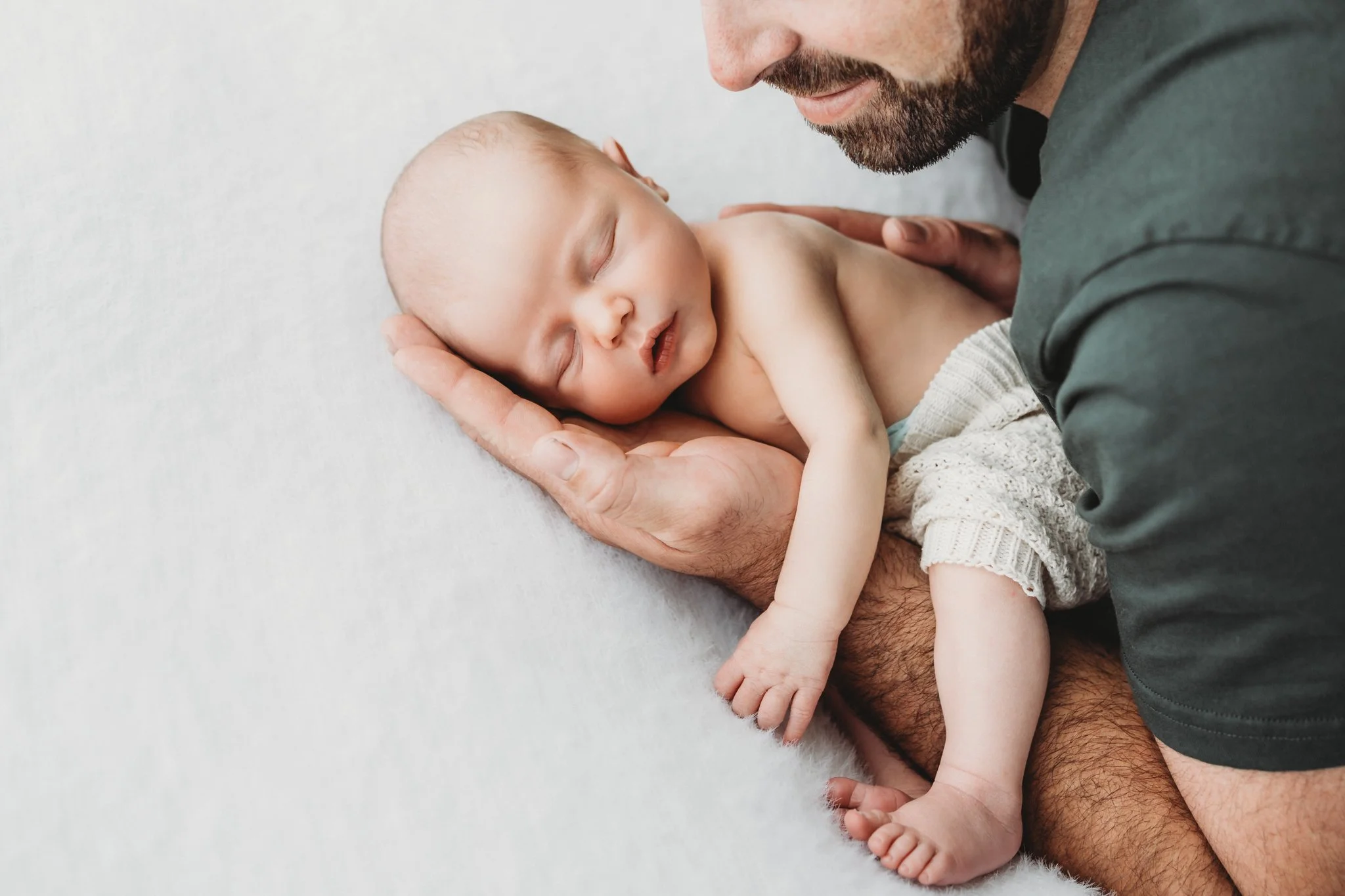 Close-up of an adult man with a beard gently holding a sleeping baby with light skin and a bald head on a soft, light-colored surface.