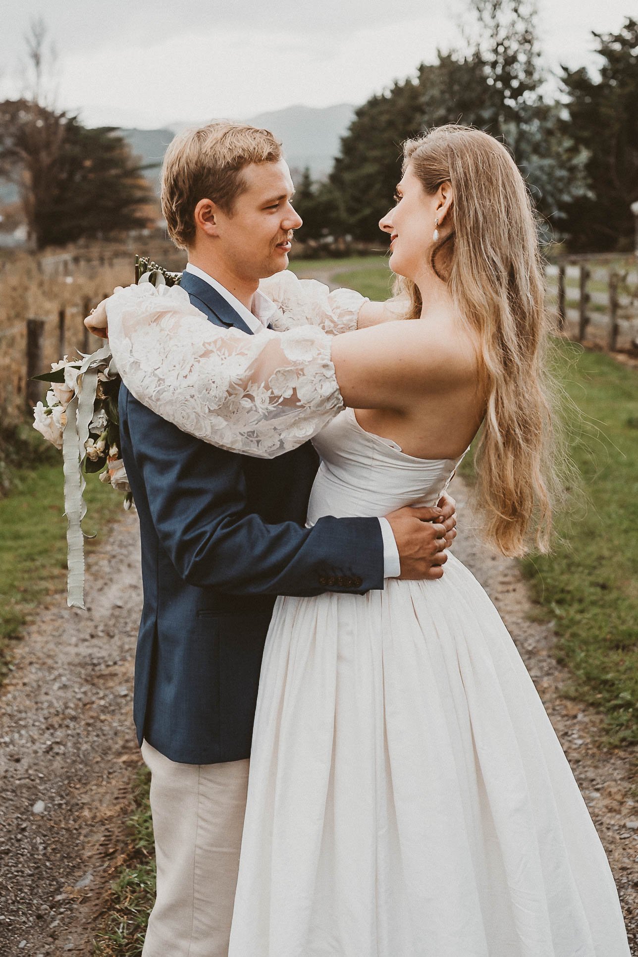 A newlywed couple sharing a tender moment outdoors, holding each other and looking into each other's eyes. The groom is wearing a navy suit with a white shirt, and the bride is wearing a strapless white wedding gown with long, wavy hair. The backgrou