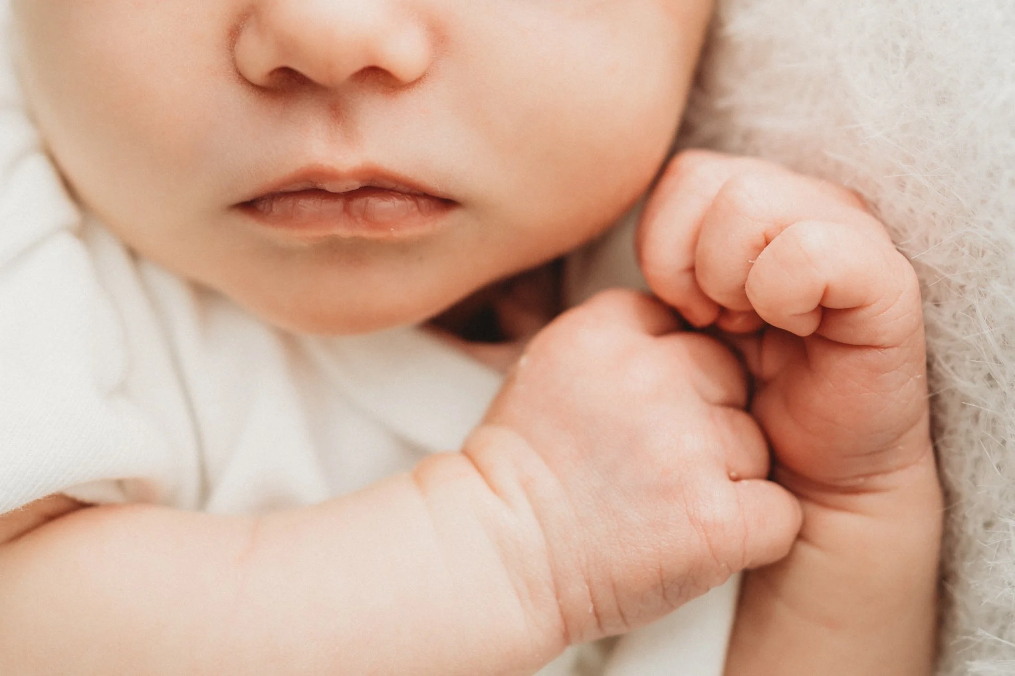 Close-up of a newborn baby's nose, lips, and a tiny hand holding an adult's finger, with soft, neutral-colored background.