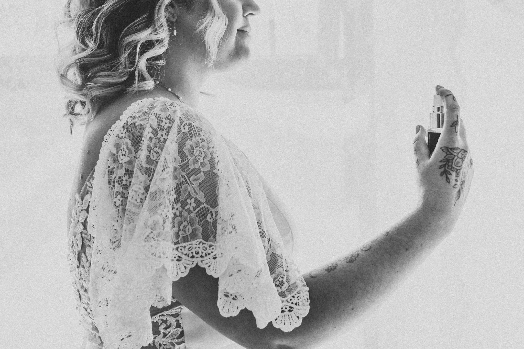 A woman with wavy hair wearing a lace robe is looking at her arm, which is being sprayed with perfume from a spray bottle held by a tattooed hand.