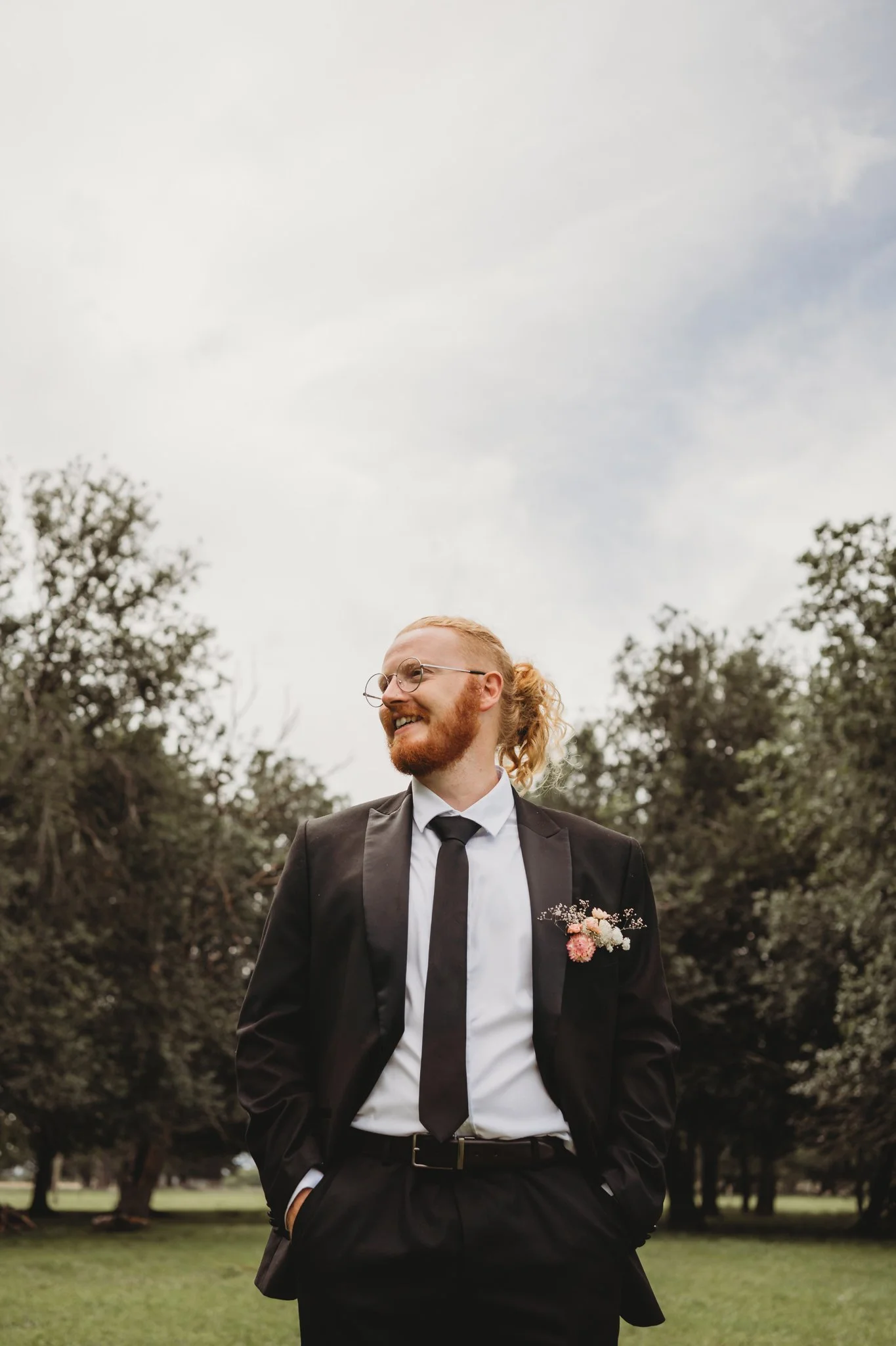 A man with red hair, a beard, and glasses, wearing a black suit, white shirt, and black tie, stands outdoors in a park with trees in the background, smiling, with hands in his pockets, and a boutonniere with pink and white flowers on his lapel.