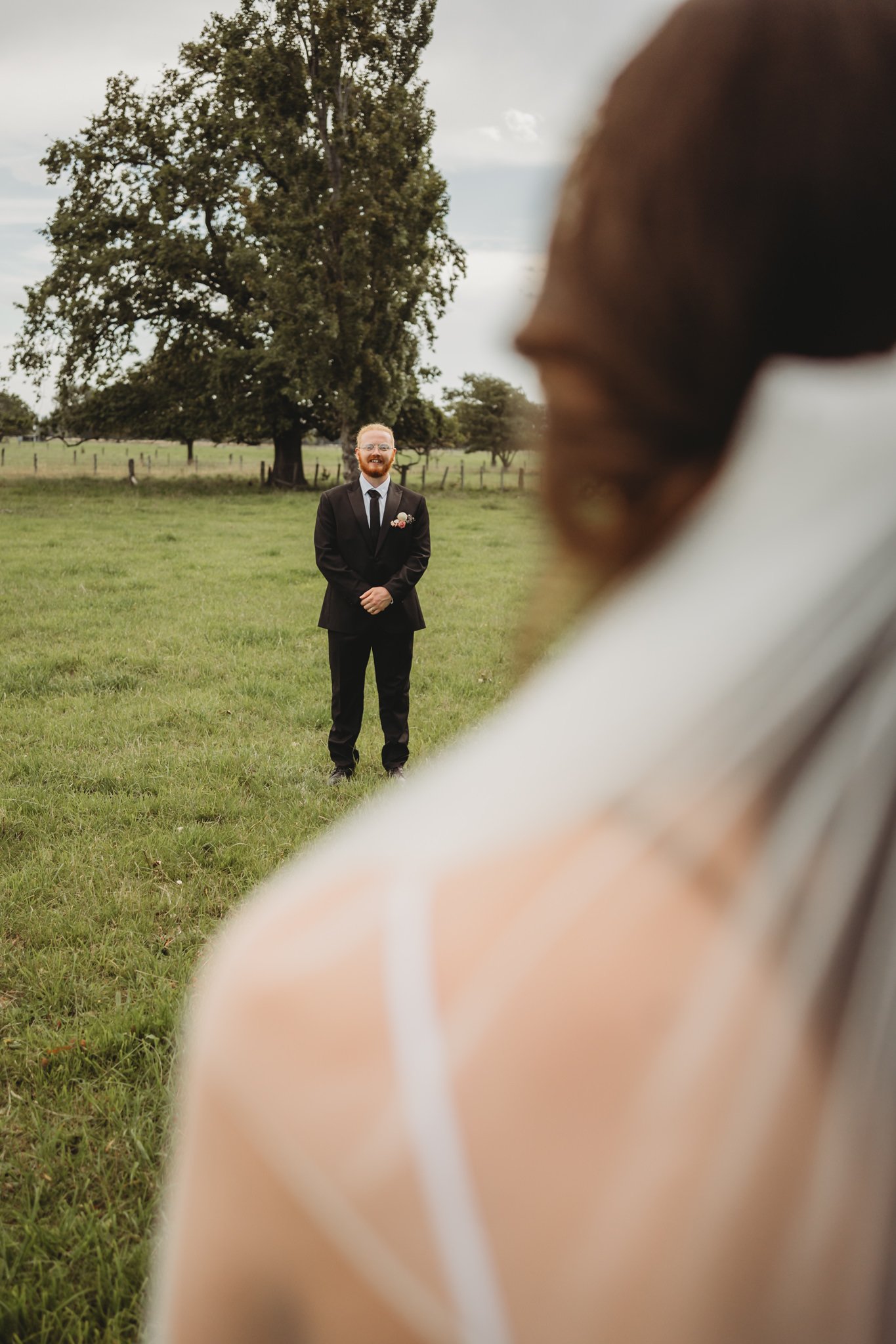 A groom in a black suit and tie, with a boutonniere, stands smiling in an open grassy field with trees and a cloudy sky. The photo is taken from a perspective that includes part of another person's shoulder and hair in the foreground.