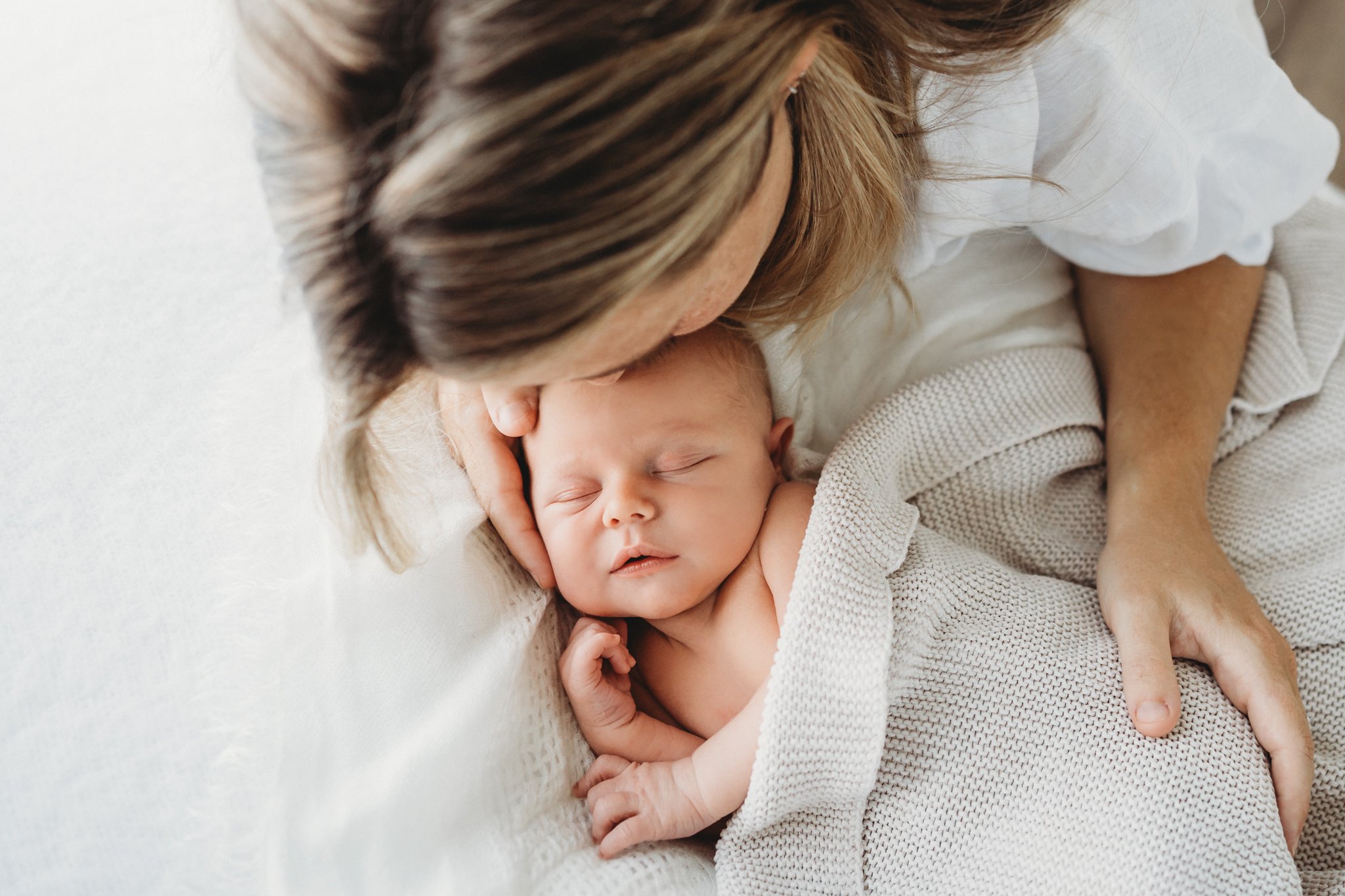 A woman with wavy hair breastfeeding a sleeping baby while lying on a white surface.