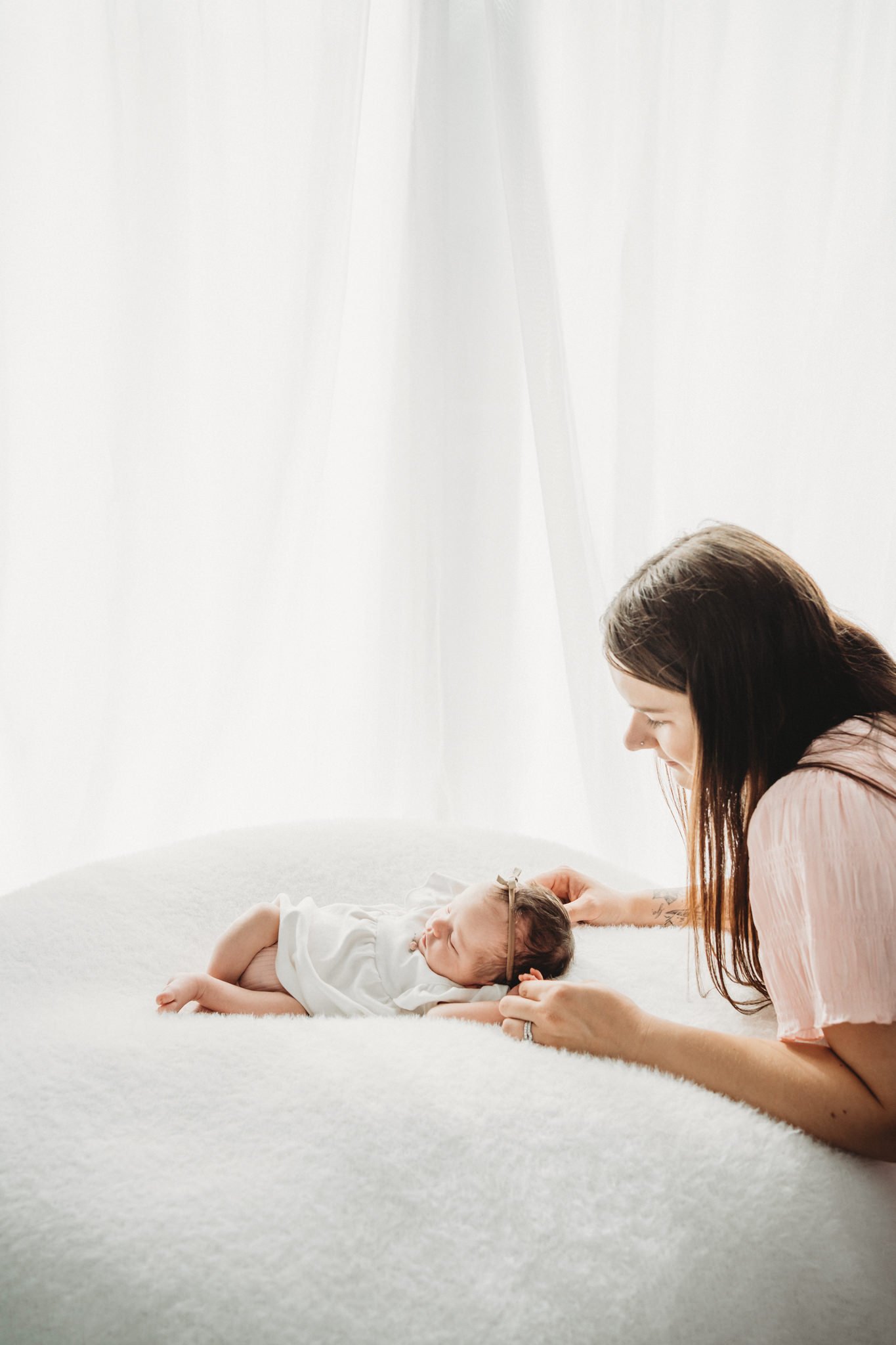 A woman with long brown hair and a nose piercing is gently holding a newborn baby wearing a white onesie while lying on a soft, white surface. The woman is leaning over the baby, appearing to interact lovingly with the infant, with a bright, minimali