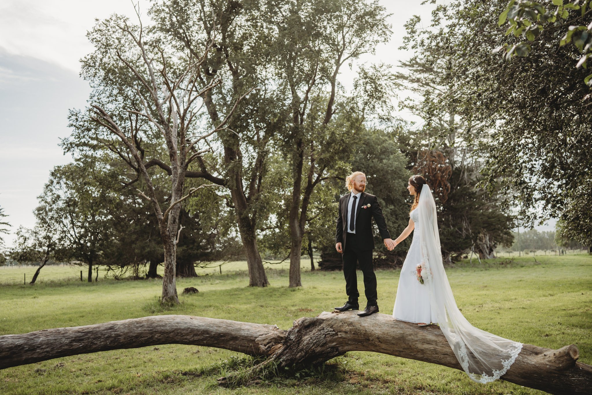 A couple dressed in wedding attire holding hands outdoors on a grassy field with trees in the background.