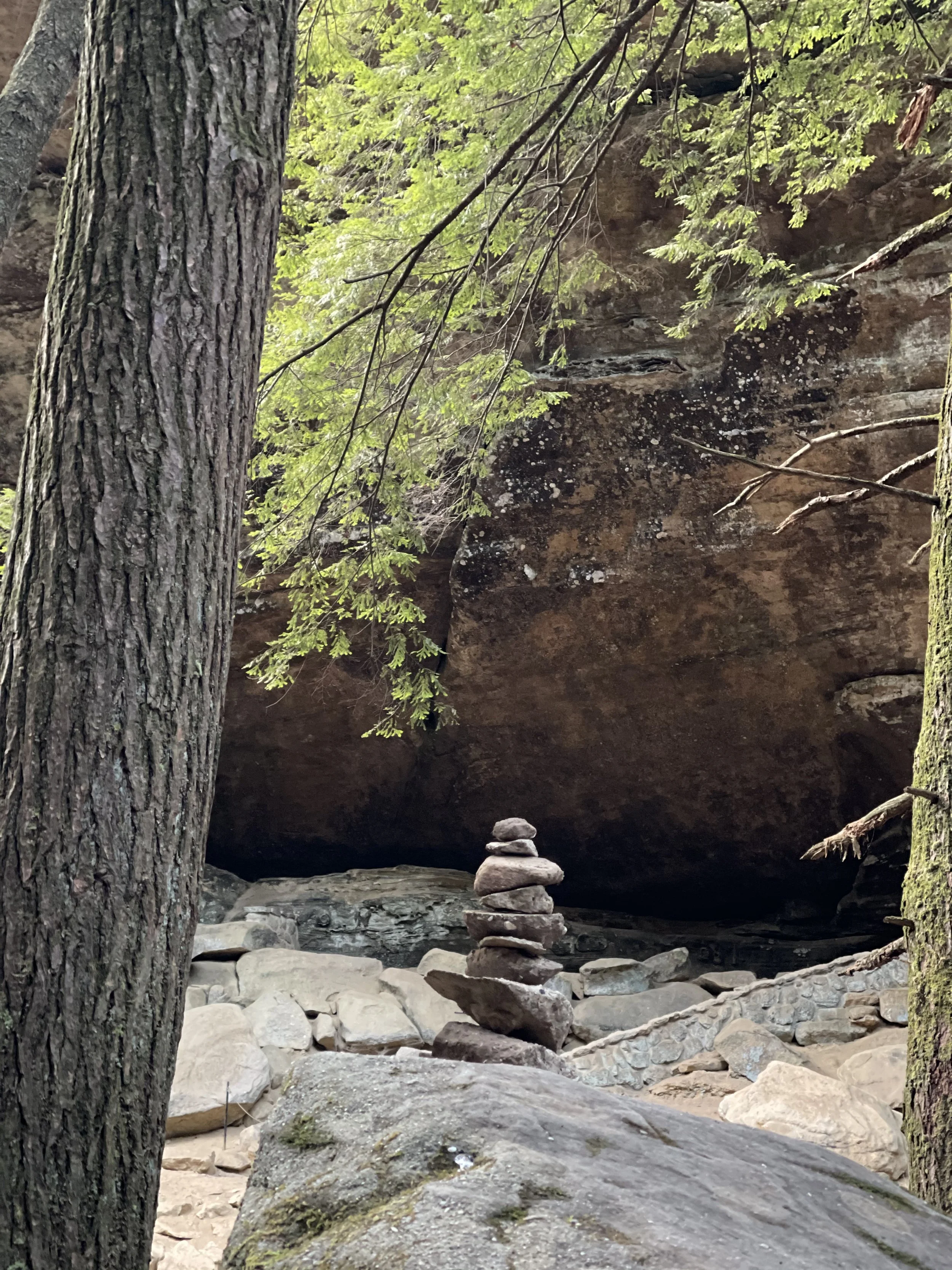 Stacked rocks forming a cairn in a forest setting with trees and a rocky area.