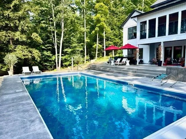 A backyard swimming pool with patio furniture and red umbrellas, surrounded by a wooded area and a modern white house with large windows.