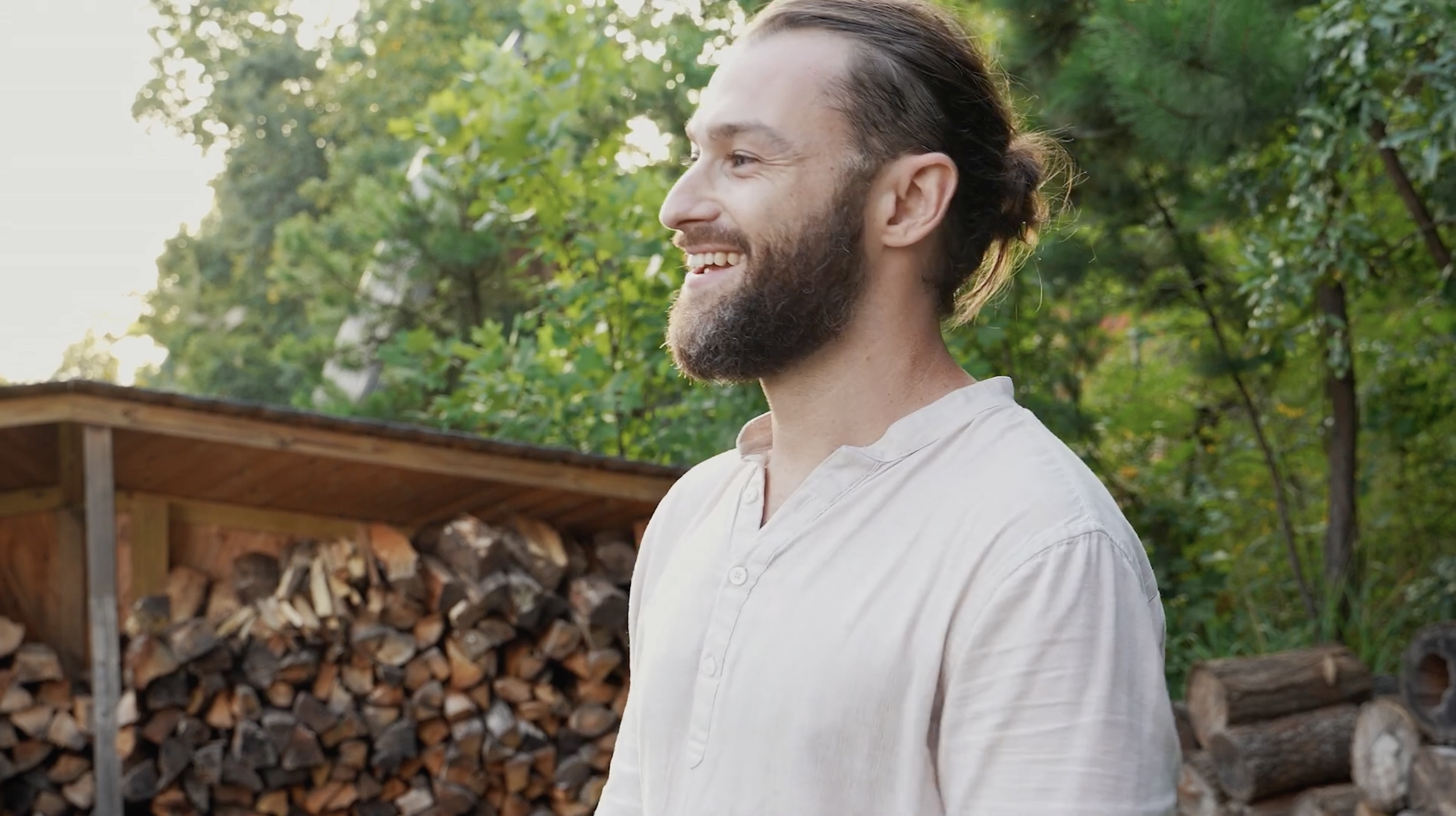 A man with a beard and long hair tied back, smiling and looking to the right, standing outdoors near a woodpile and green trees.