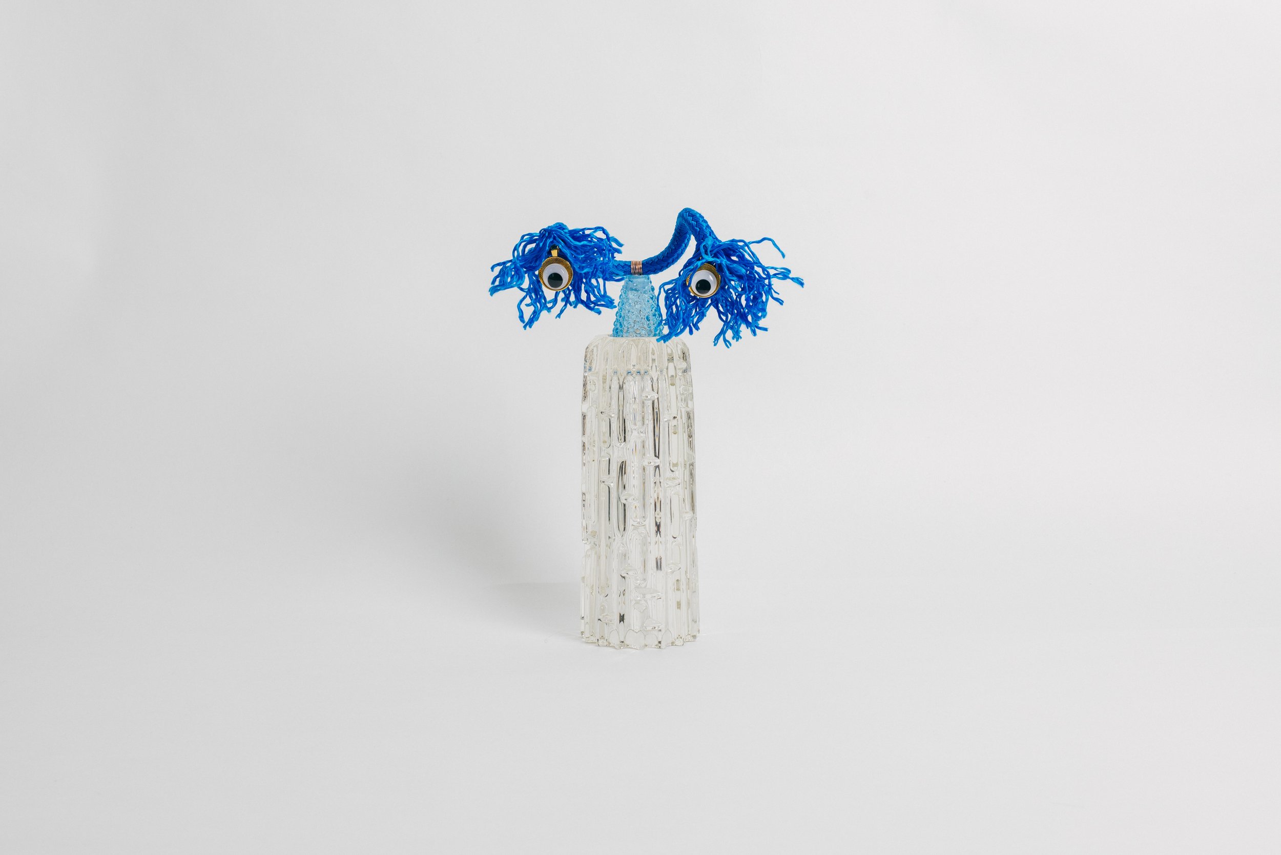 A glass sculpture resembling a face with blue yarn hair, googly eyes, and a blue nose against a plain white background.