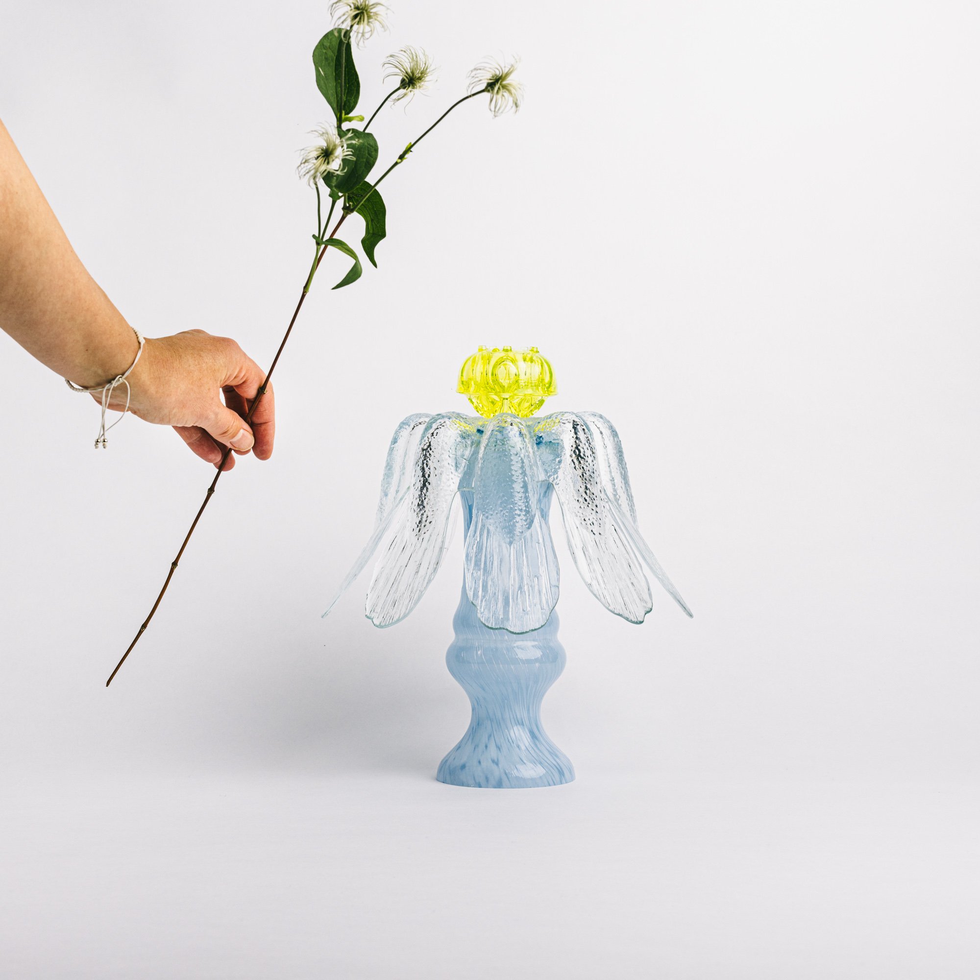 A hand holding a flower with green leaves and white flowers, positioned above a glass vase. The second chance sculptural art design resembling an angel with wings, made of clear and light blue glass, with a yellow decorative element on top.