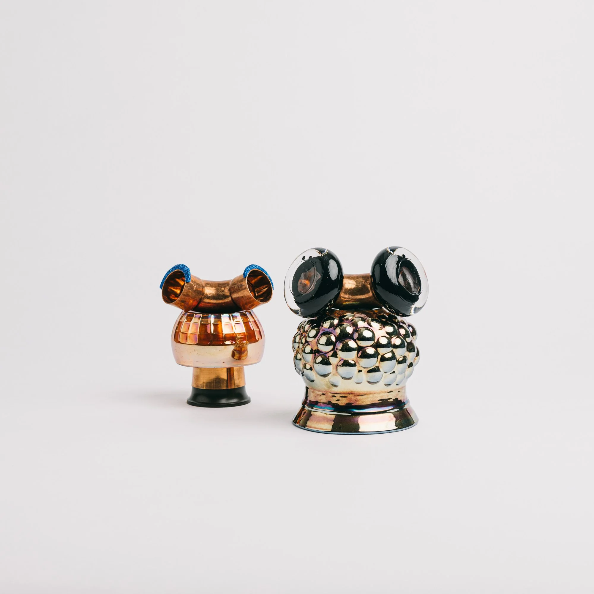 Repurposed materials created siru tuomisto second chance art these whimsical Two shiny, metallic, abstract sculptures with rounded shapes and eye-like elements, set against a plain white background.