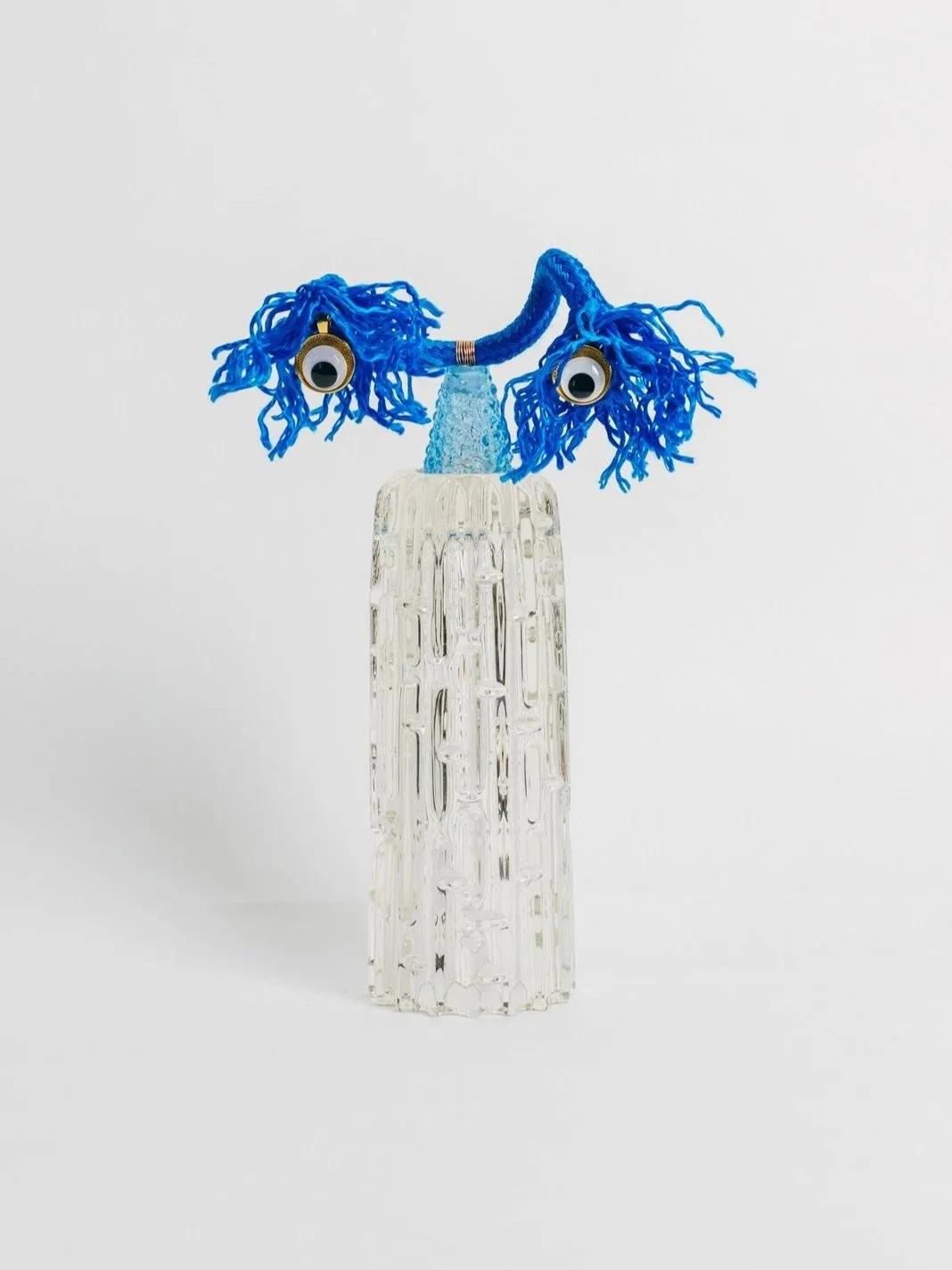 A glass art sculpture Enlightened Iceman resembling a face with large googly eyes and blue yarn hair against a plain white background.