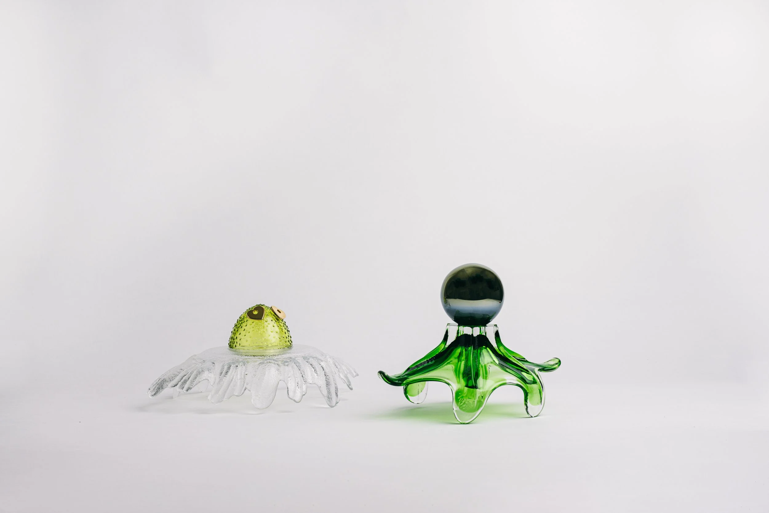 Two glass art sculptures resembling sea creatures on a white background, one green with a black sphere on top and the other white with yellow eyes, both with flowing shapes.