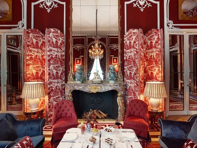 The Santo Mauro Hotel's red dining room has red wallpaper and an ornate fireplace with an elegant mirror reflecting an impressive chandelier. The table is set with cocktails and appetizers before the start of a Christmas Day lunch.