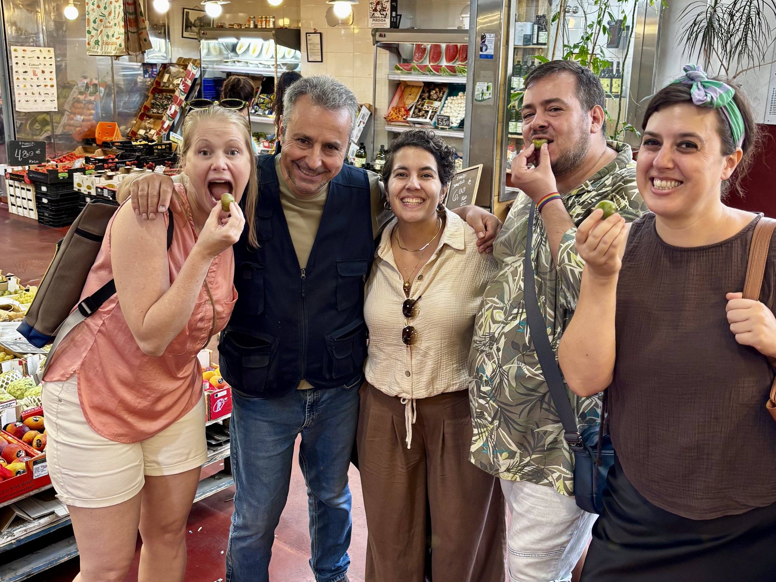 A group of four people bite into seasonal fruit alongside the fruit vendor, Pedro, during a visit to a traditional market in Madrid.