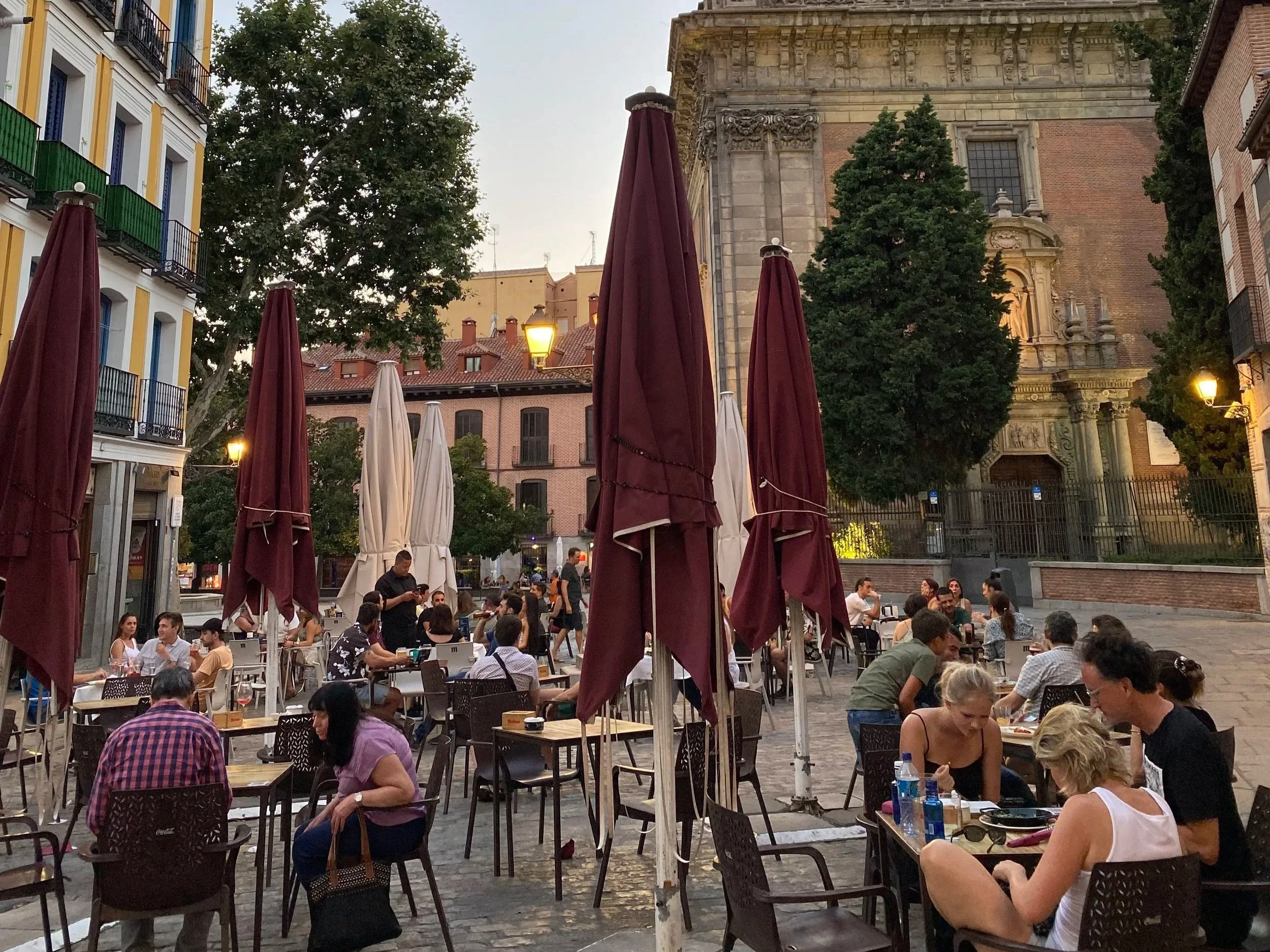 Tables fill a plaza in Madrid at sundown as families enjoy food and drinks.