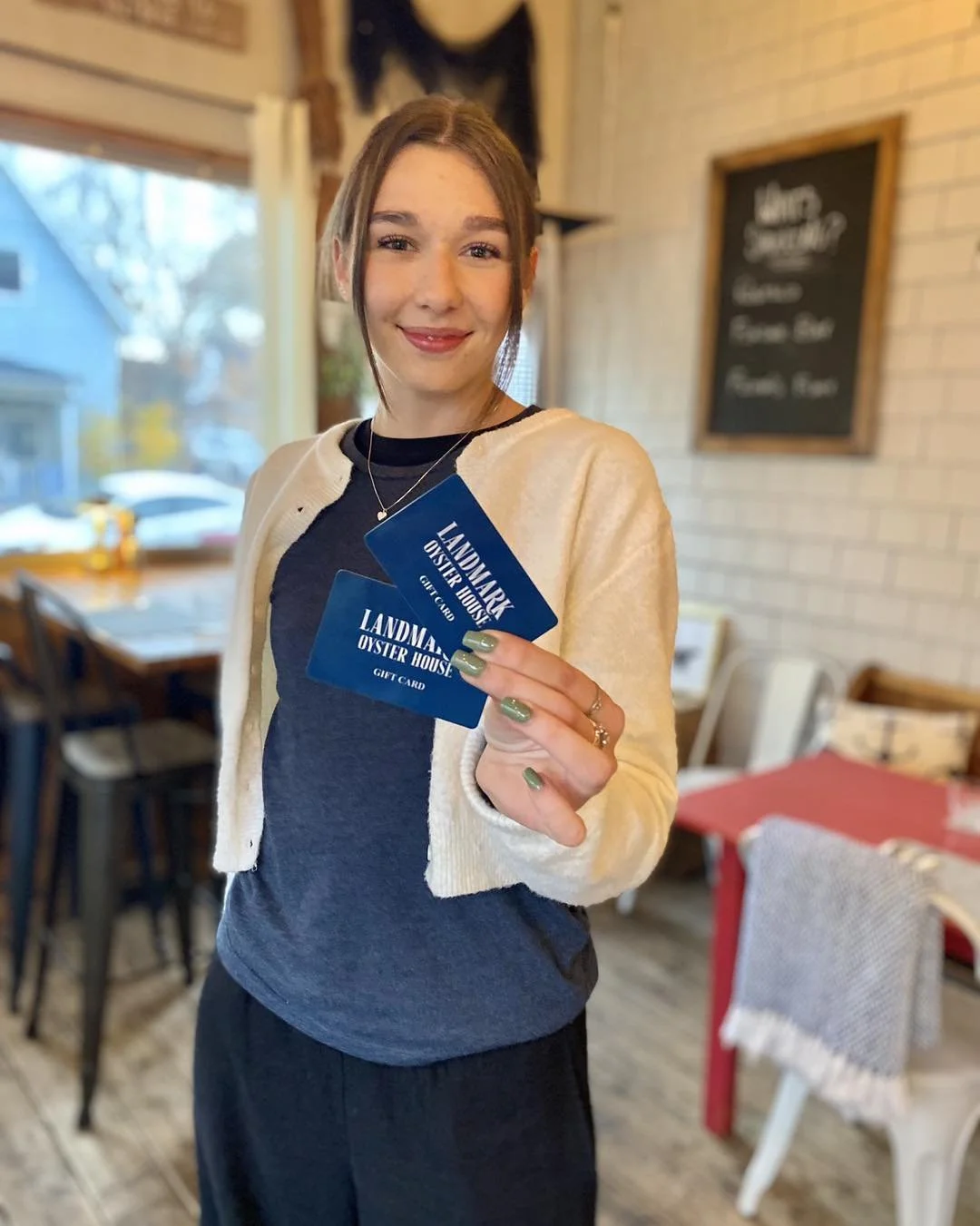Young employee in white sweater and blue Landmark Pyster House branded t-shirt holds up two gift cards