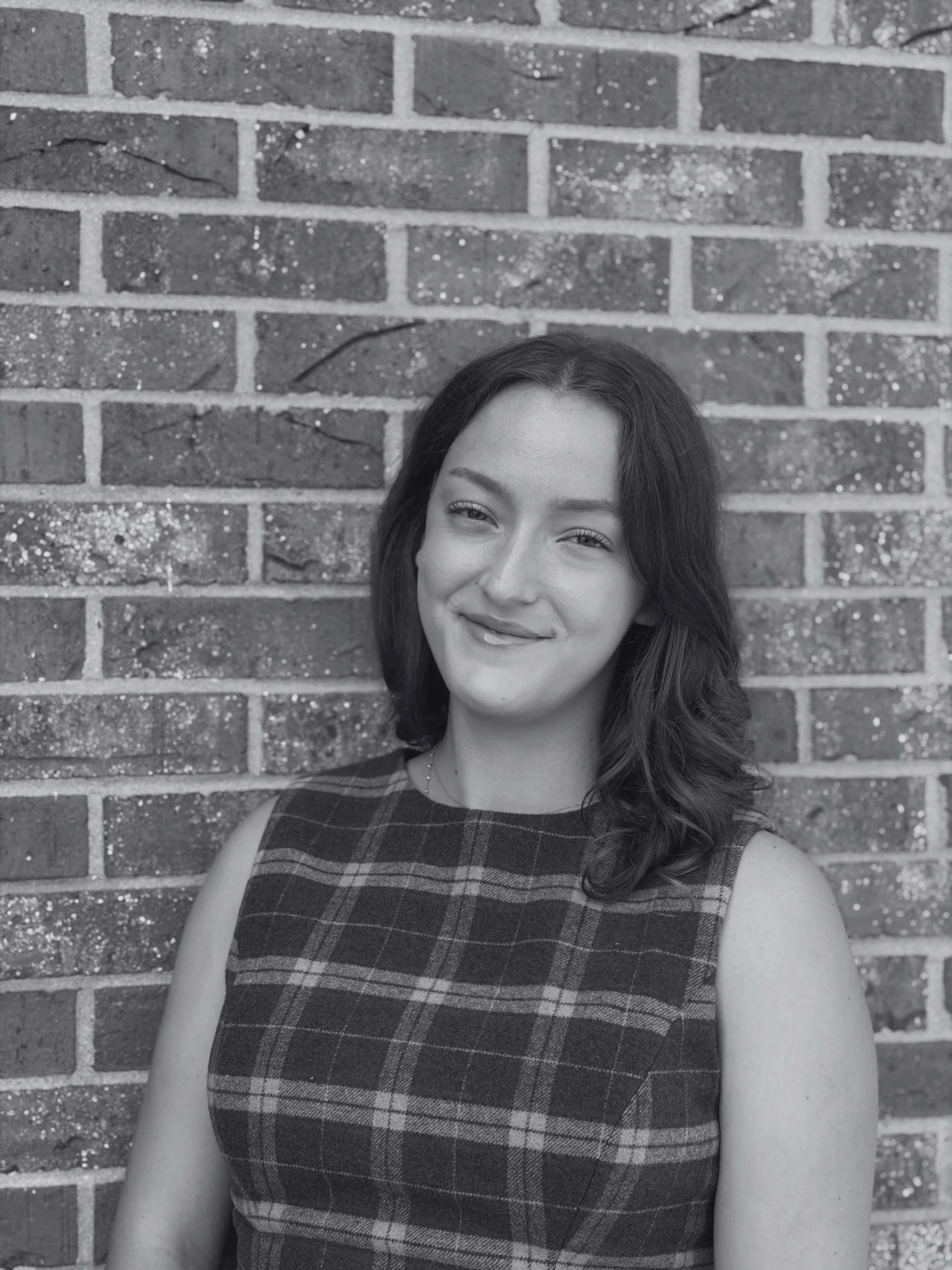 A woman with long dark hair and light skin smiling, standing against a brick wall, wearing a sleeveless plaid dress.