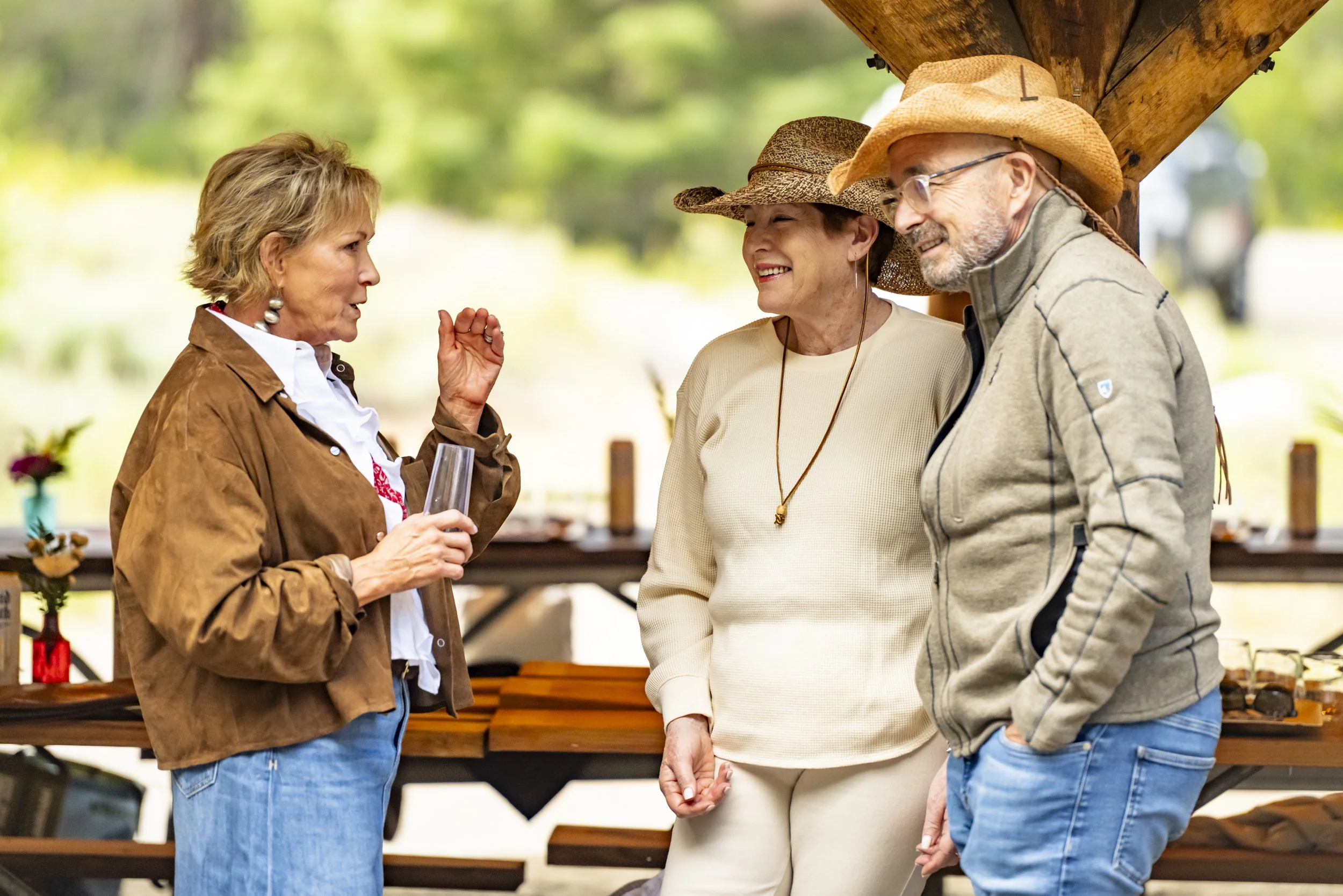 Three older adults engaged in conversation outdoors under a wooden structure, with picnic tables and trees in the background.