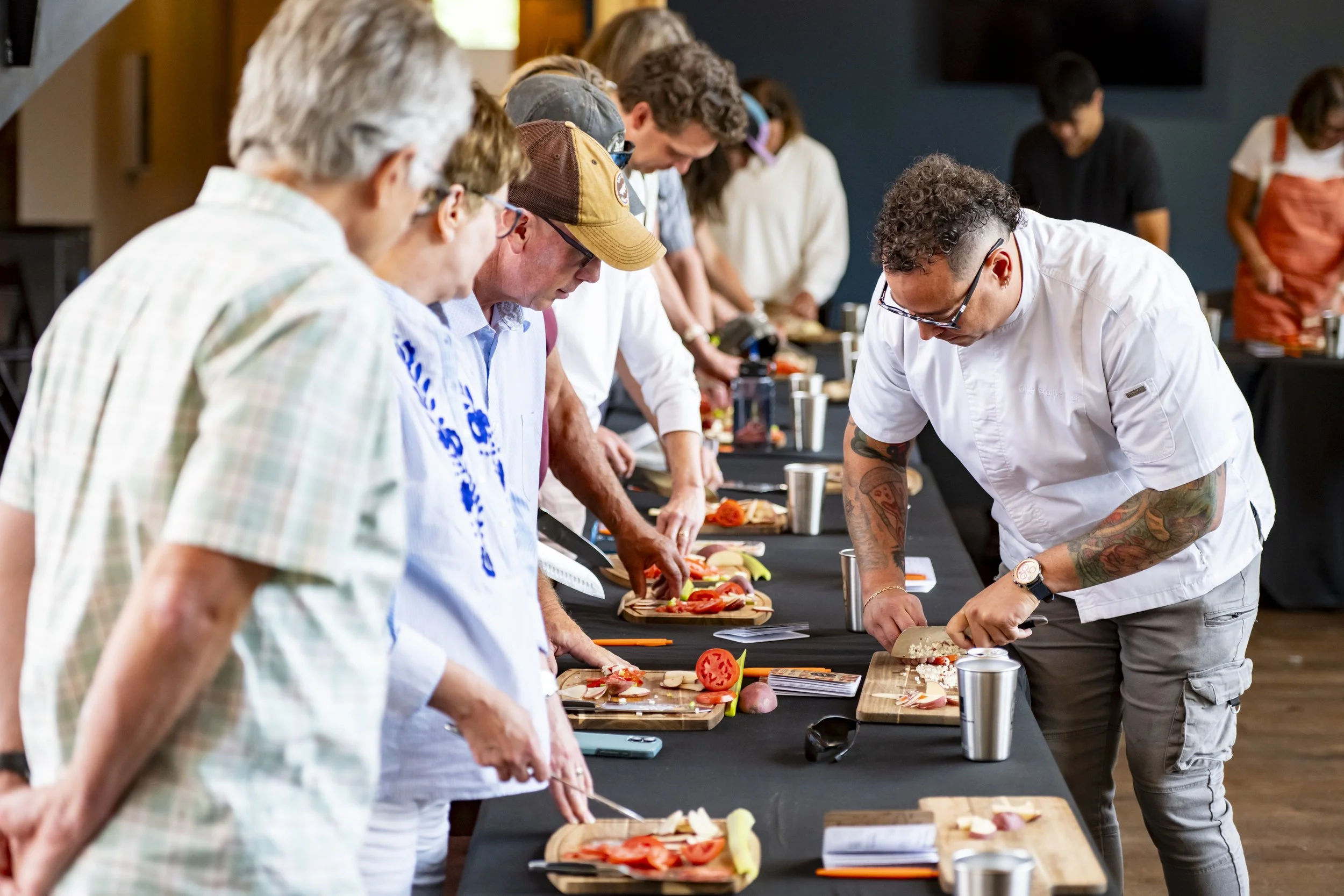 A group of people participating in a cooking class, preparing food on cutting boards at a long table.