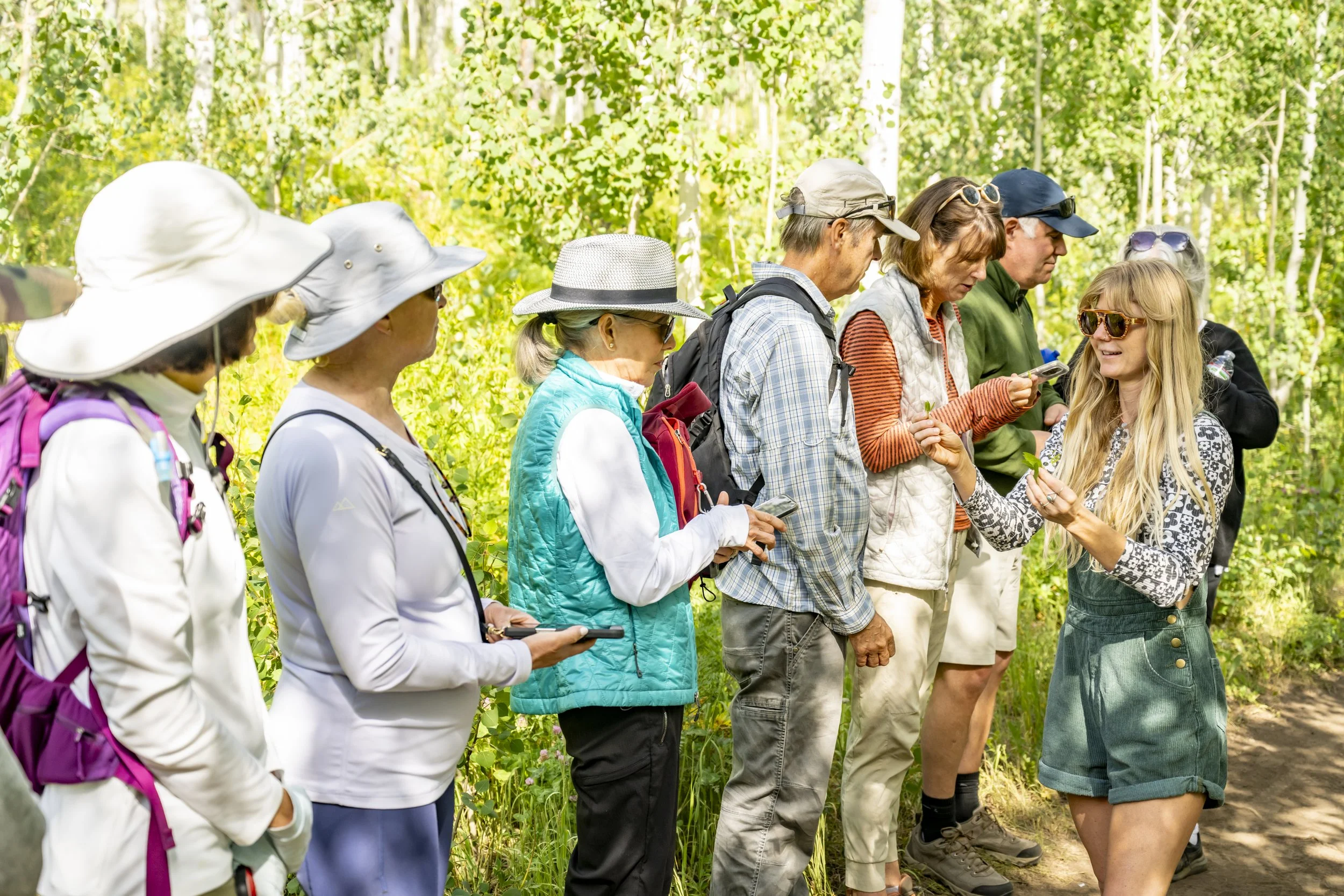 Group of people on a nature hike, standing in a line in a forest with green trees, some using smartphones, and a woman at the front demonstrating with a plant.