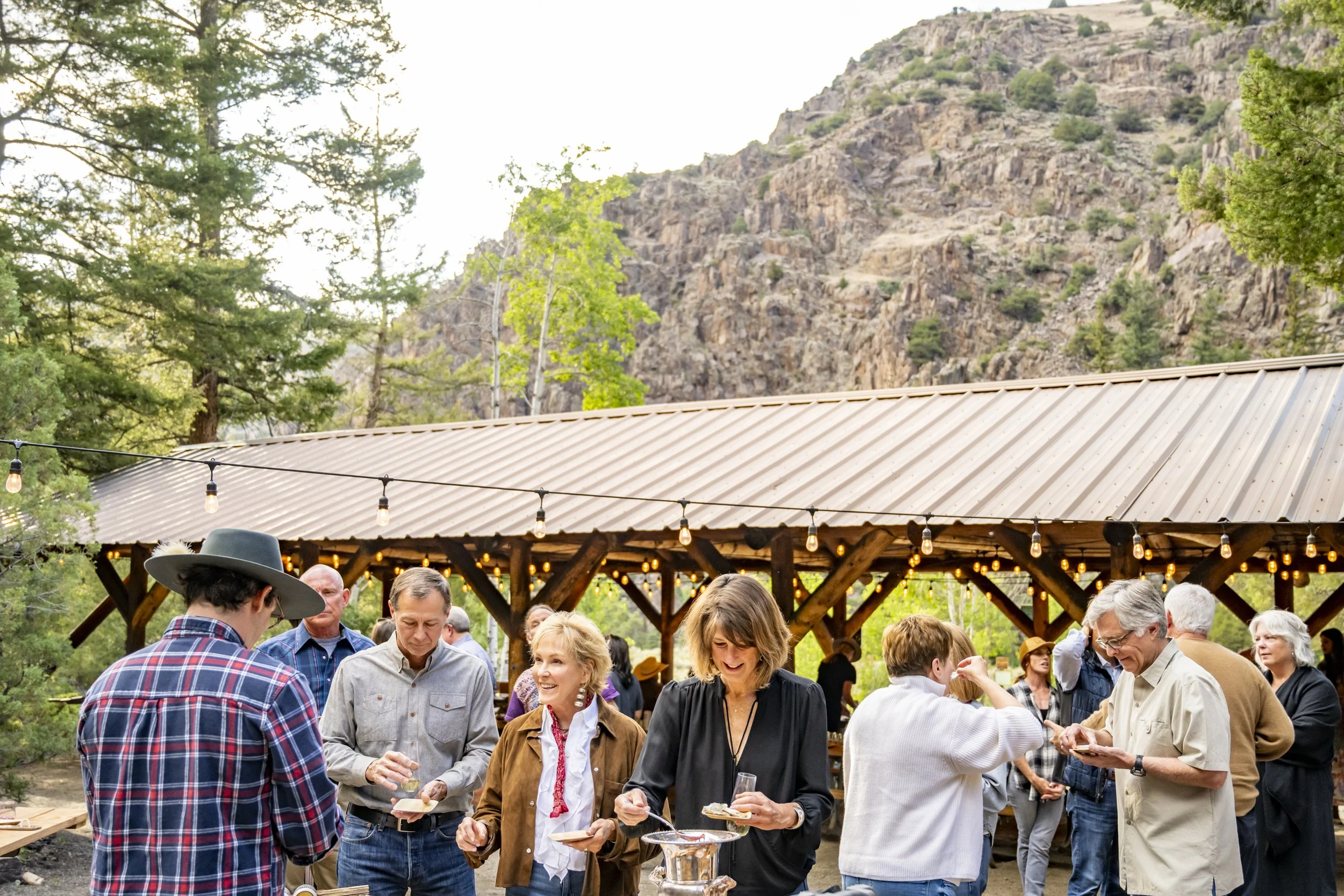 Group of people enjoying outdoor gathering under string lights with a mountain backdrop.