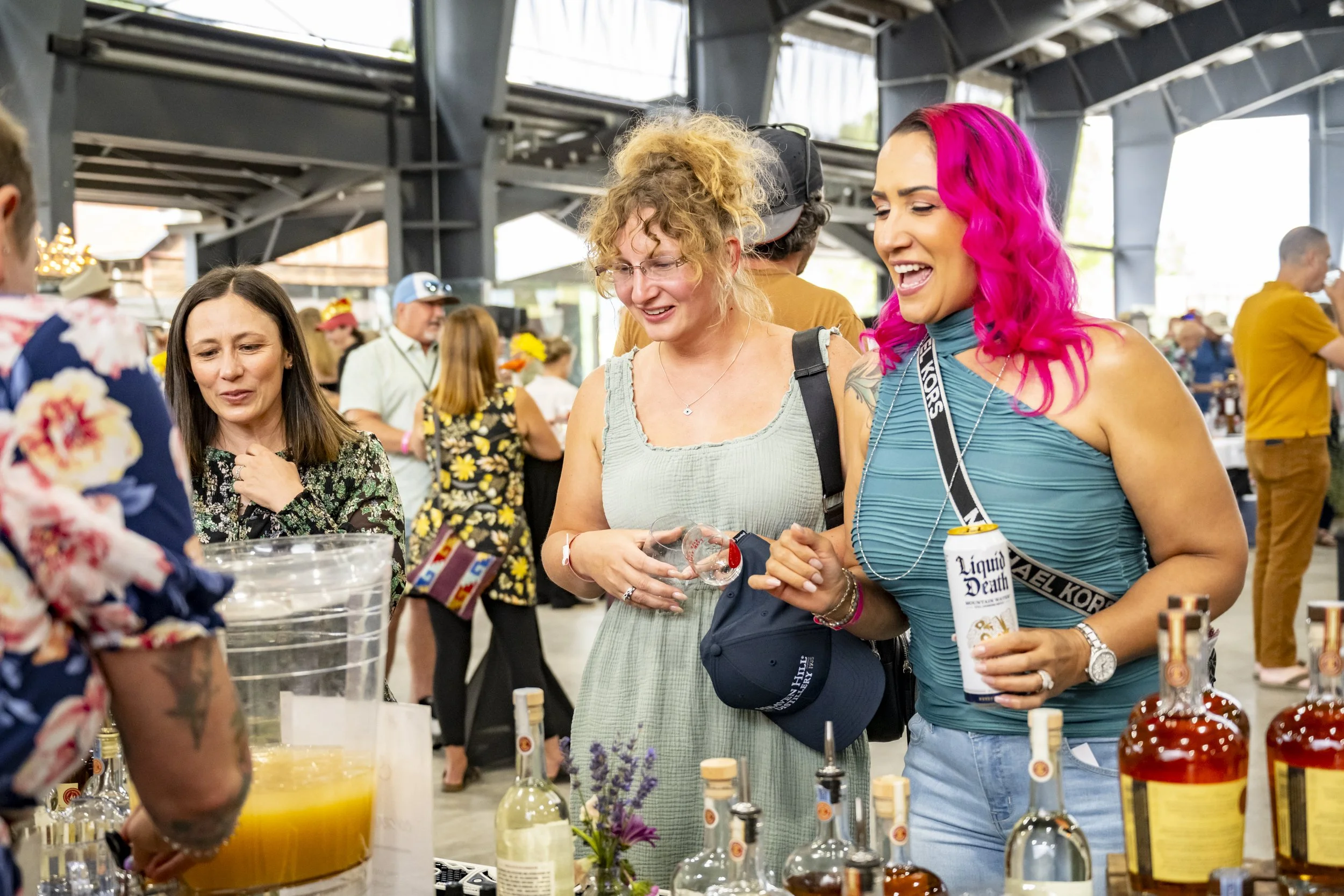 Three women at a beverage booth, smiling and talking, with bottles and mixers on the table, in a crowded indoor event space.