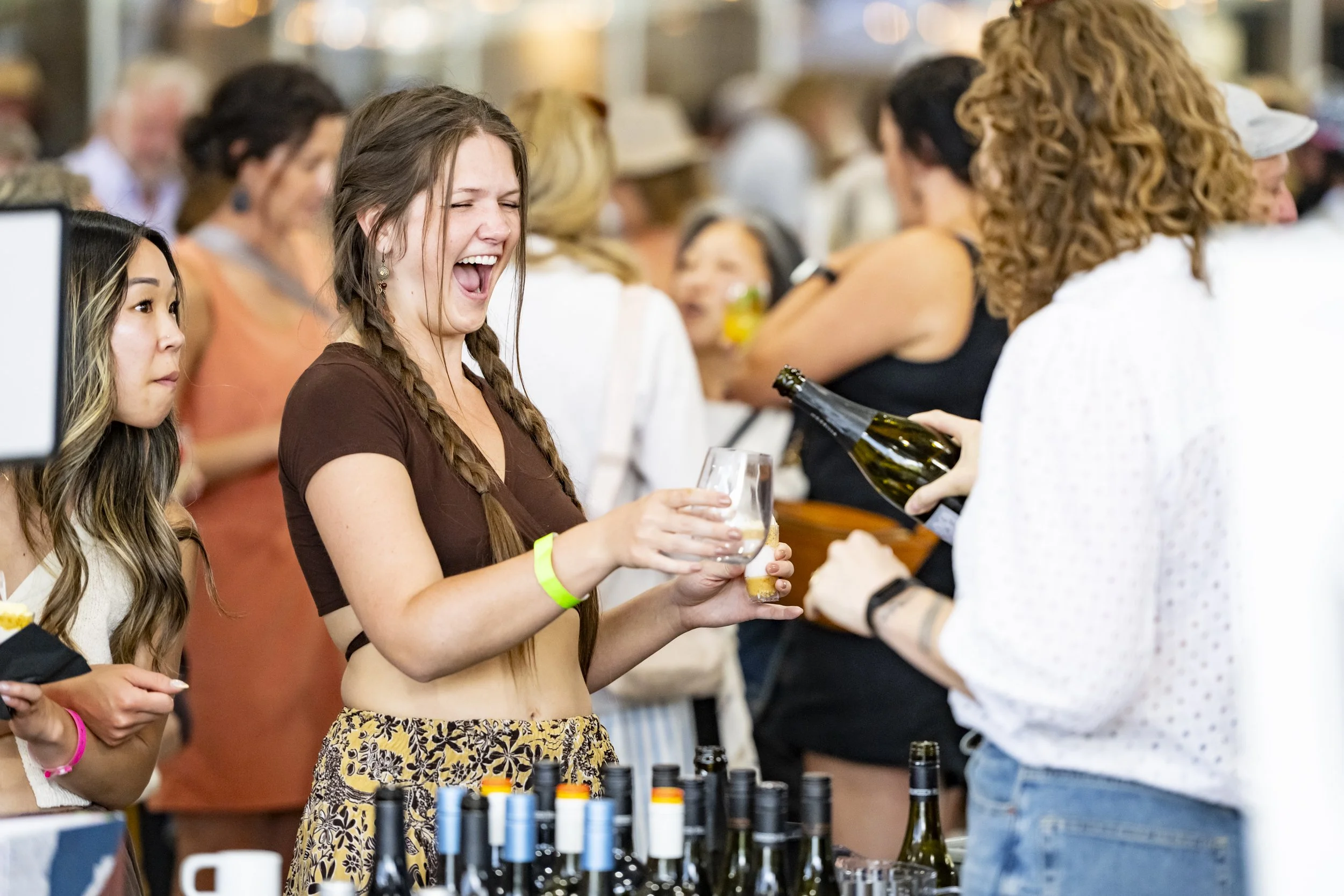 Woman with braided hair and a brown crop top laughing while holding a glass, as another woman pours her a drink at a crowded social event.