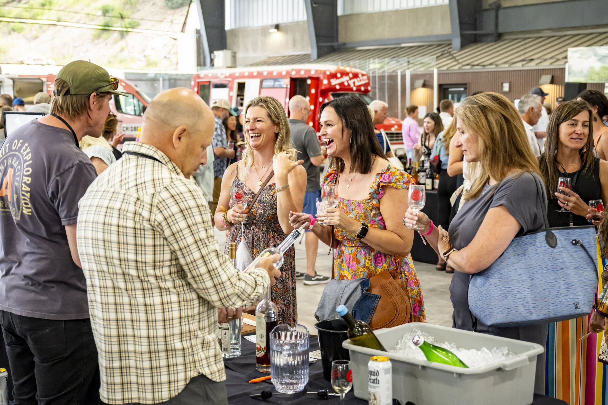 People socializing at a tasting event, with a man pouring drinks at a table and women holding glasses, smiling and laughing.
