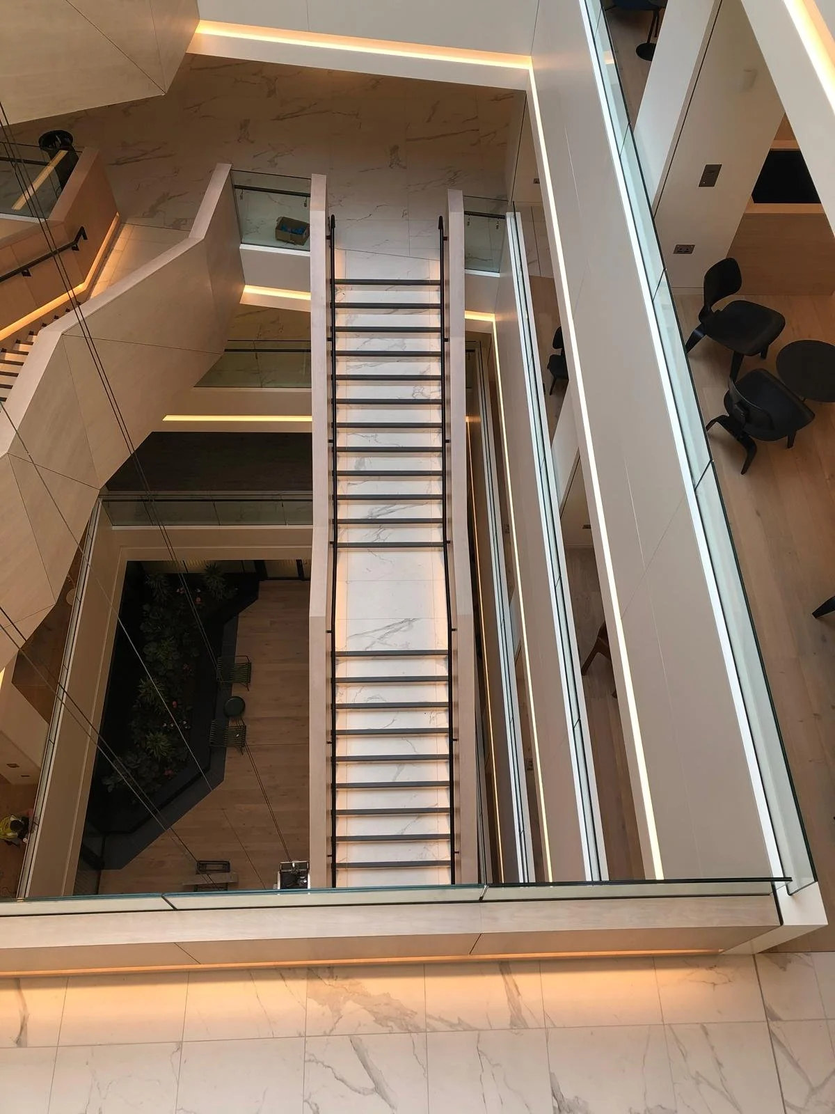 Modern multi-story interior with staircase and glass railings, viewed from above, showing marble flooring, black chairs, and hallway with lighting accents.