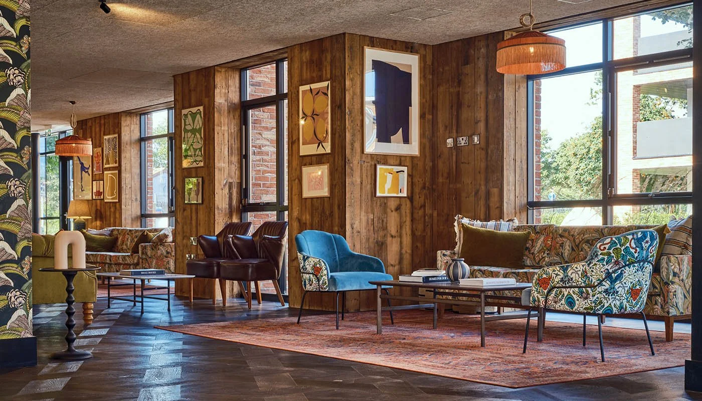 Living room with large windows, wooden paneled walls, colorful upholstered chairs, patterned sofa, coffee tables, artwork on the walls, a rug, and natural light streaming in.