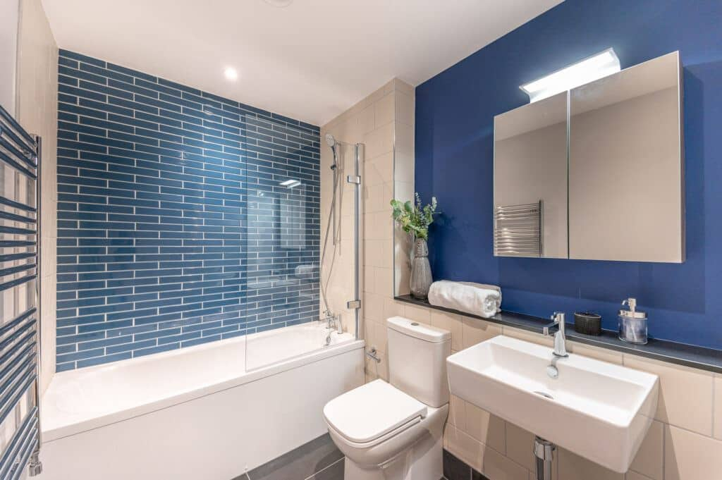 Modern bathroom with blue accent wall, white bathtub with glass shower screen, toilet, white sink with faucet, black countertop, and a mirror cabinet. Decorative vase with flowers and bathroom essentials on countertop.