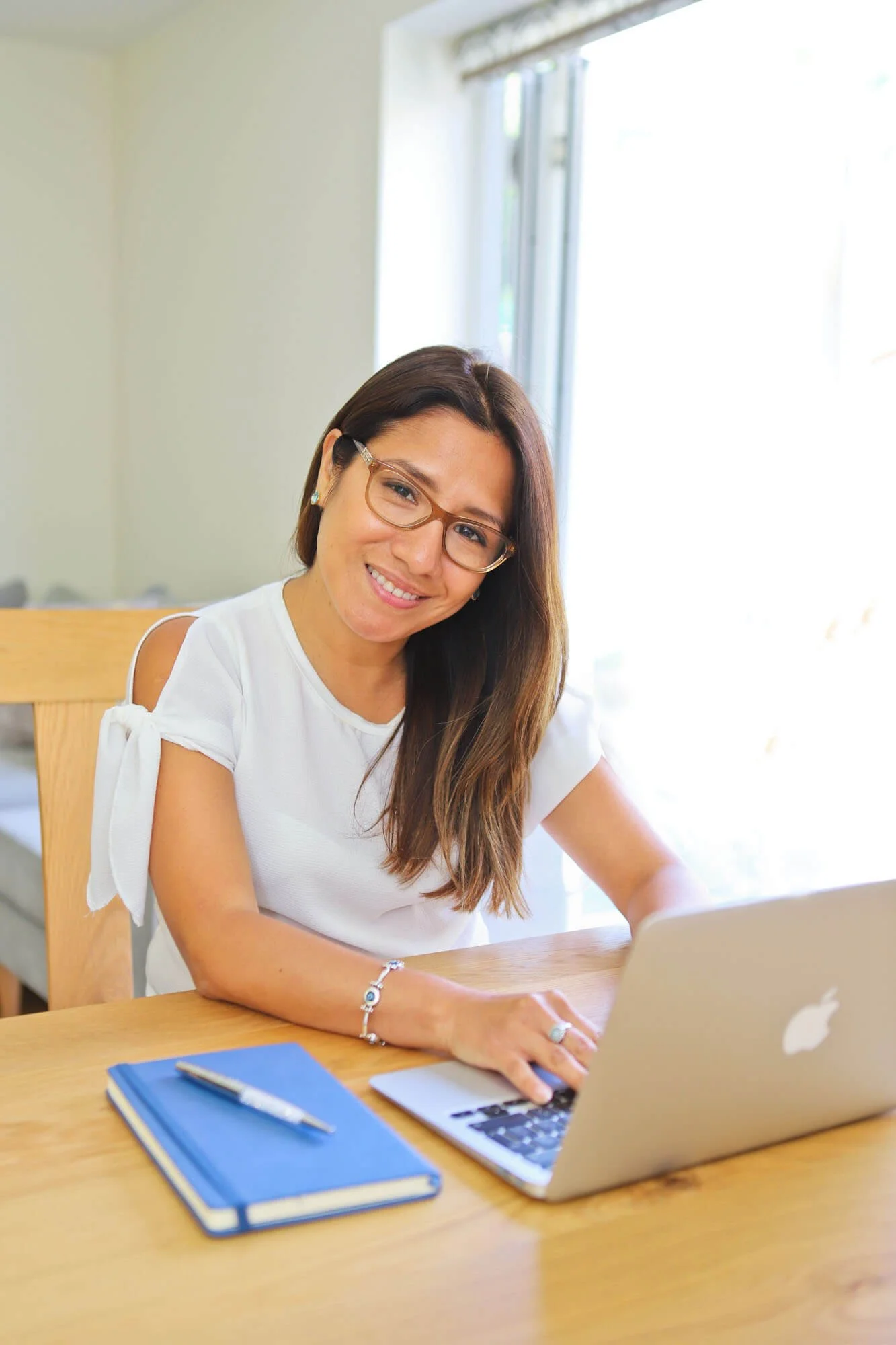 Lucía sitting at a wooden table working on a laptop. There is a blue notebook, a pen, and a smartphone on the table. She is smiling and in a well-lit room with a large window behind her.