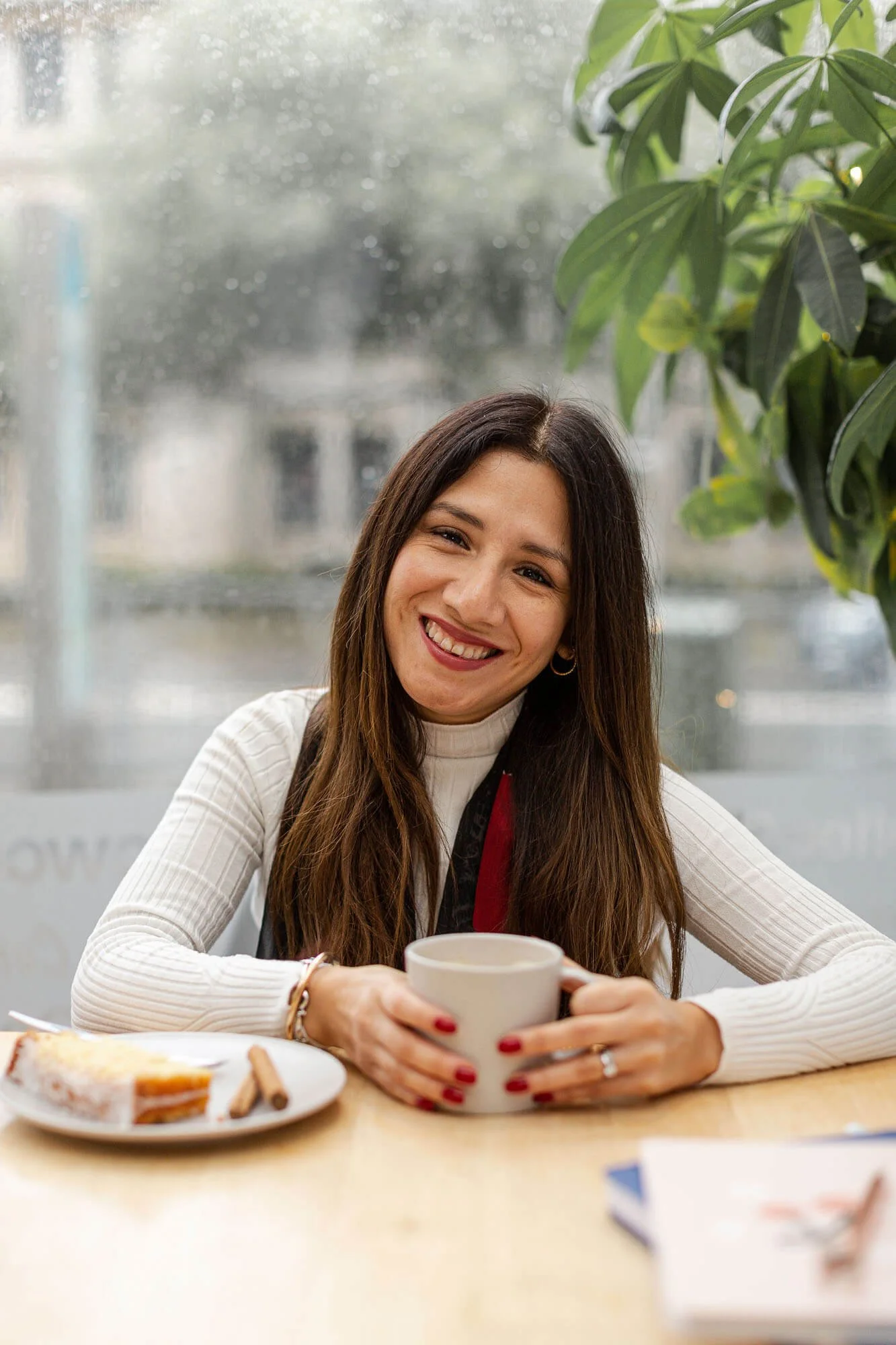Lucía smiling while holding a mug, sitting at a table with a breakfast plate in front, in a room with a window and green leafy plant in the background.