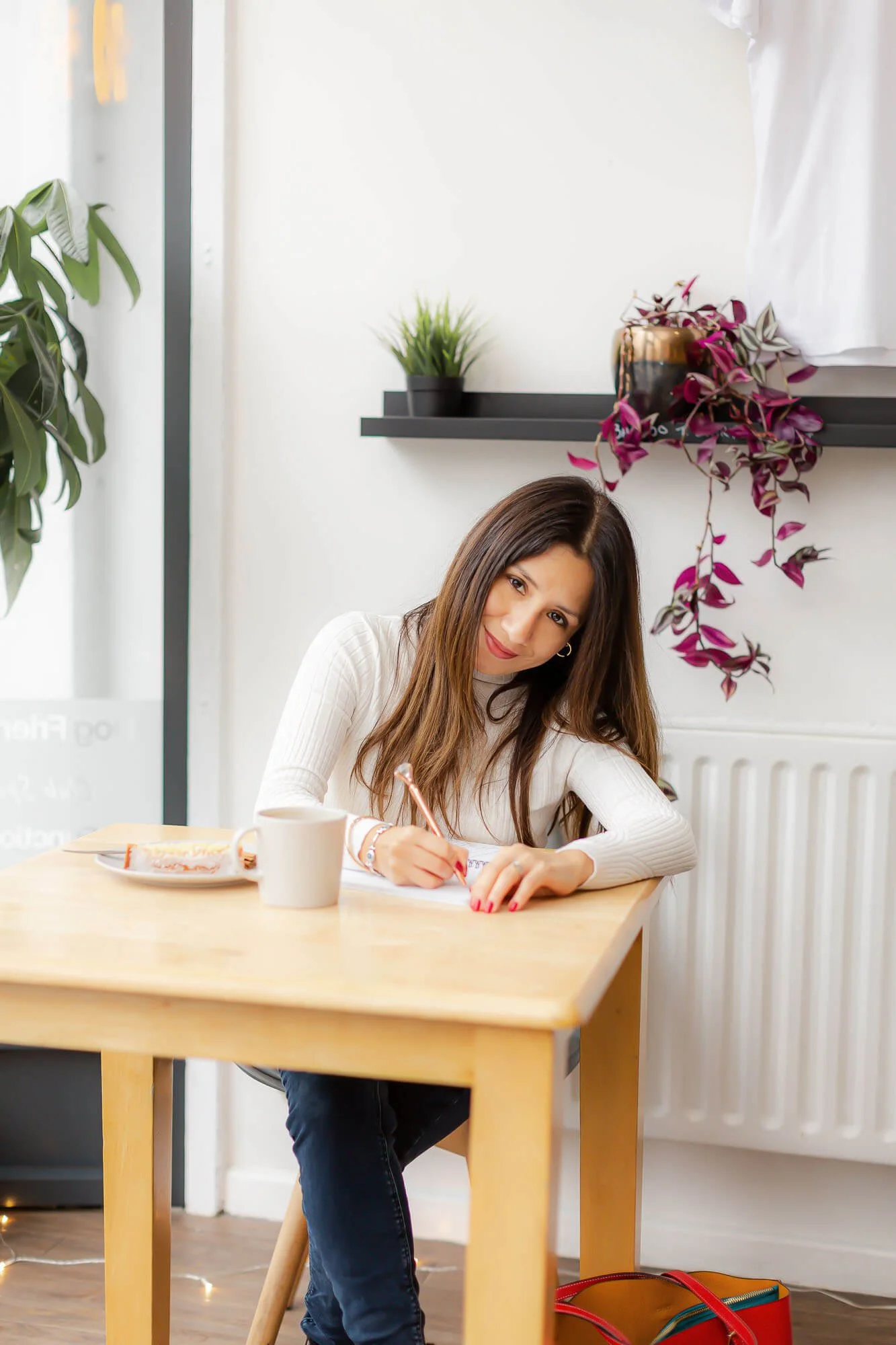 Lucía sitting at a wooden table, writing in a notebook with a mug and a plate of pastries in front of her. The room has white walls, a black shelf with green and purple plants, and light coming from a large window.