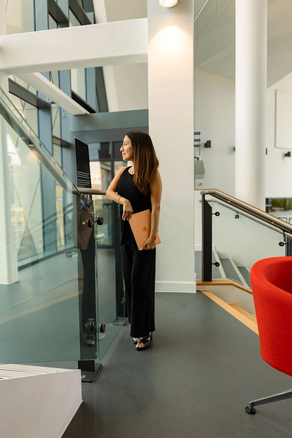 Lucía holding a closed rose gold laptop, standing inside a modern office building near a glass railing, with a staircase and large windows in the background.