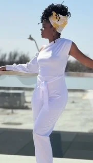 A woman in a white jumpsuit standing outdoors near a railing, wearing sunglasses and a yellow face mask, with her arms outstretched.