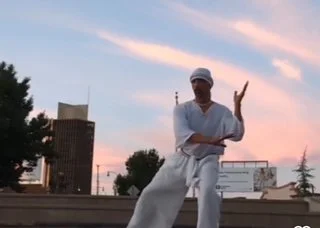 Person dressed in white clothing and a cap performing a martial arts move outdoors during sunset with city buildings in the background.